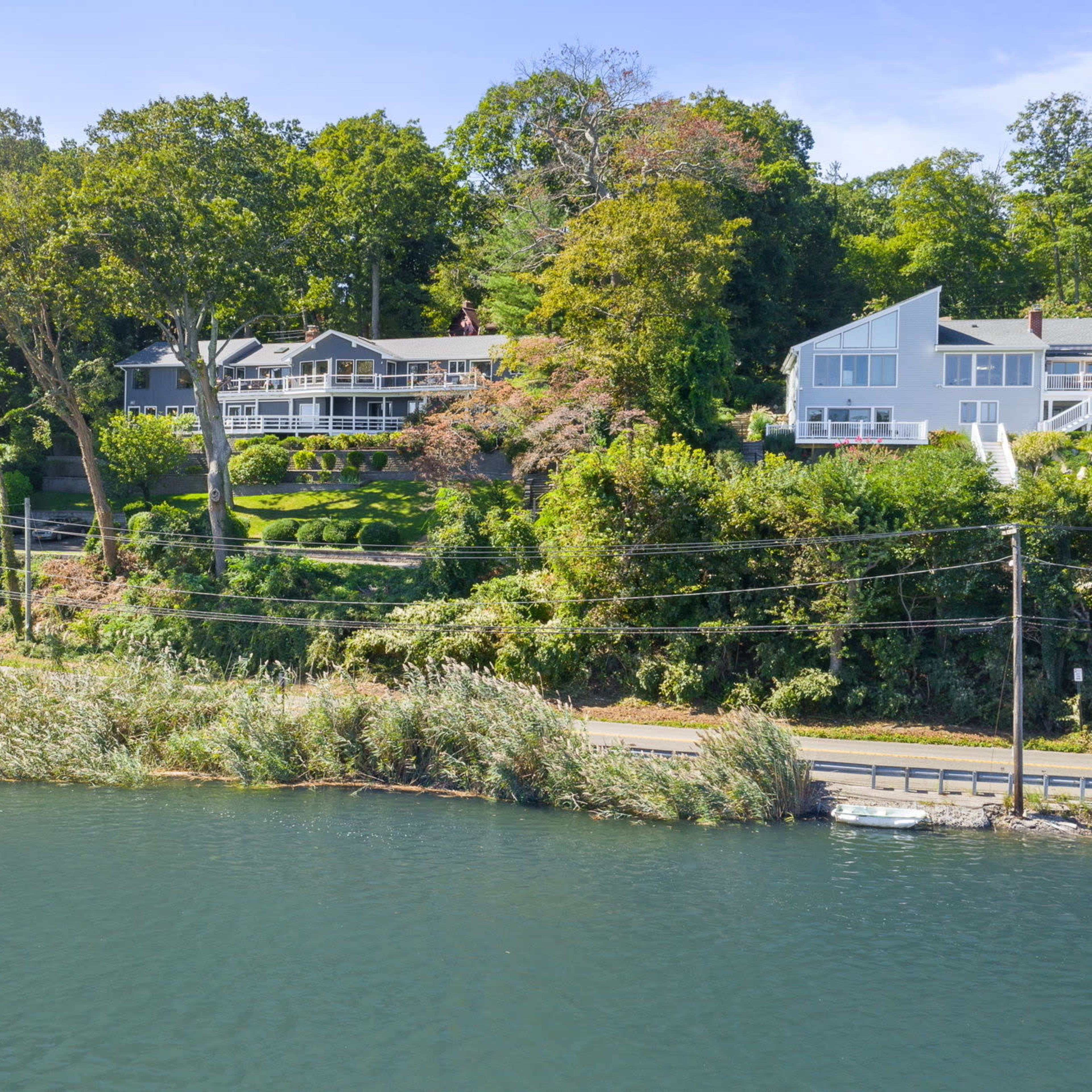 A row of houses is situated on a hillside above a calm river, with lush trees lining the bank and power lines running alongside the road below.