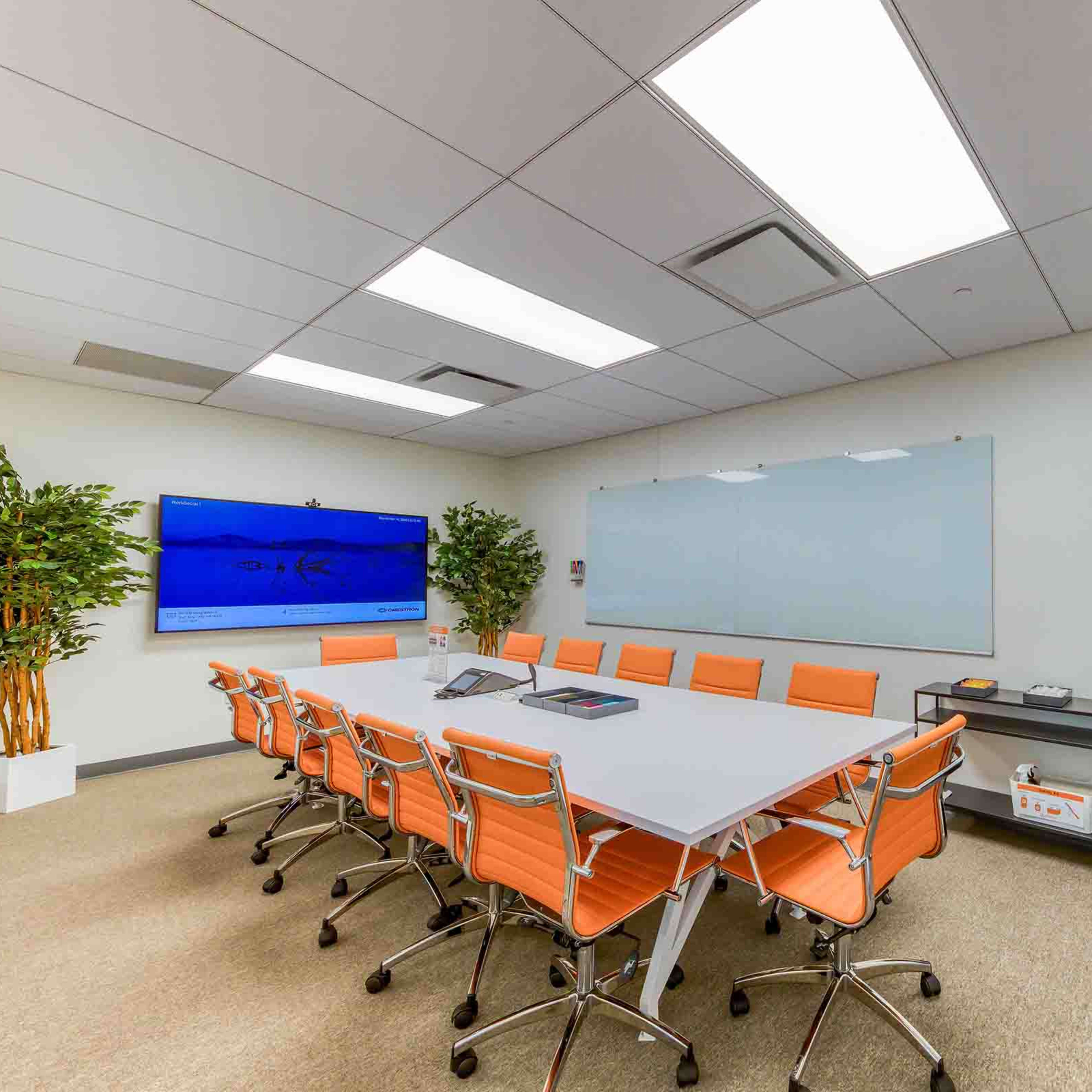 A brightly lit conference room features a large table surrounded by orange chairs, a glass whiteboard, and a display screen on the wall.