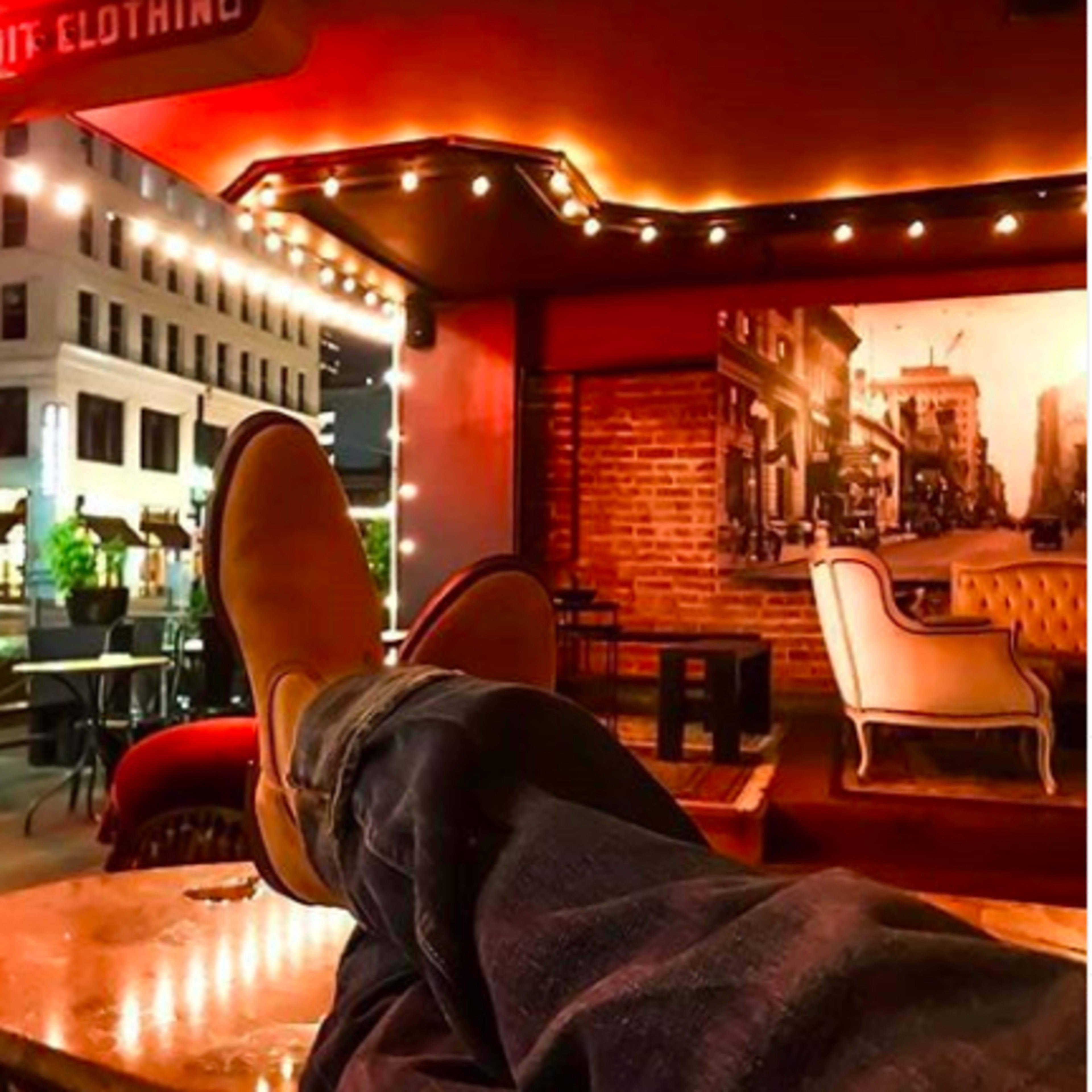 A person relaxes with their feet up on a table in an outdoor seating area, surrounded by twinkling lights and city buildings at night.
