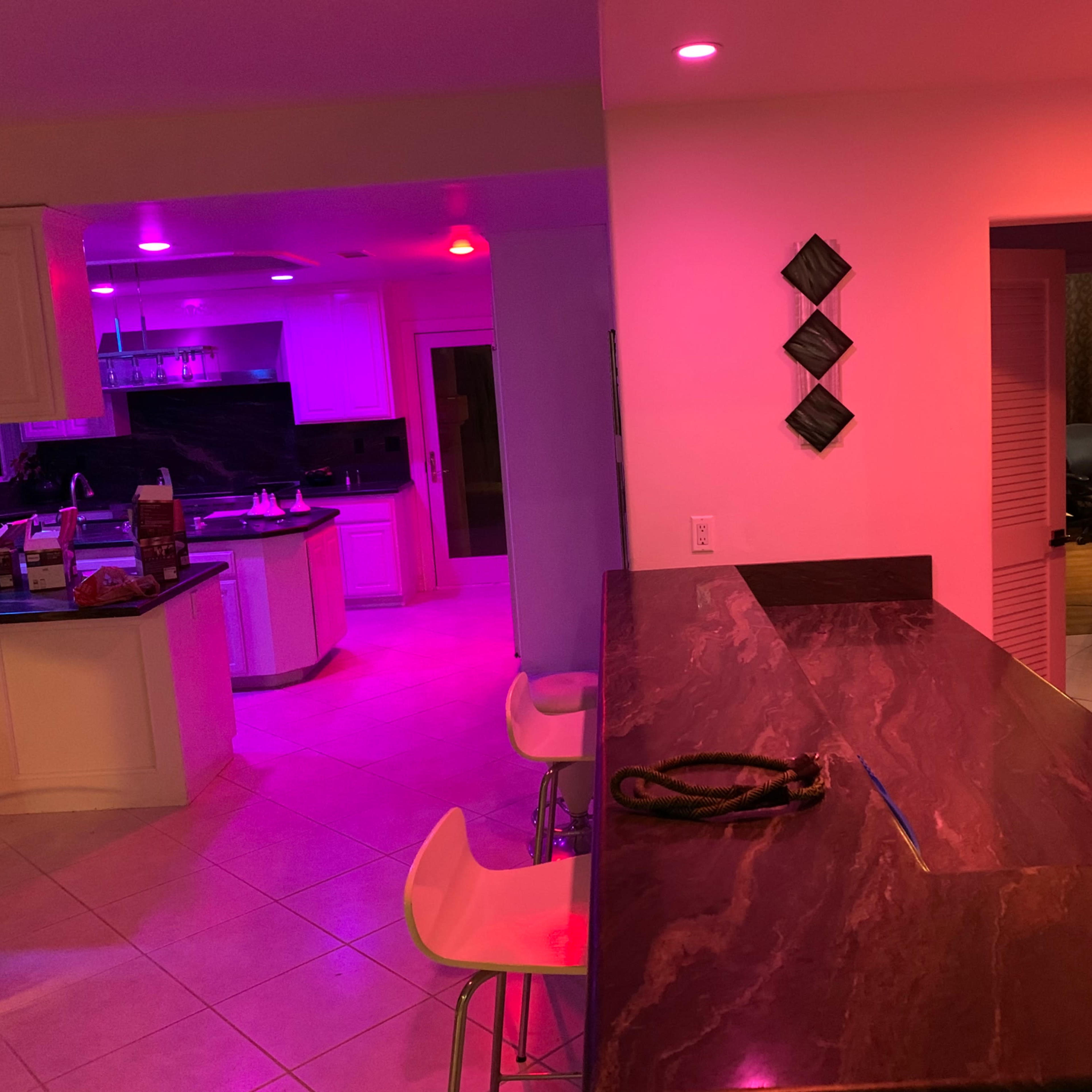 A modern kitchen and bar area illuminated with pink and purple lighting, featuring a marble countertop and white cabinetry.