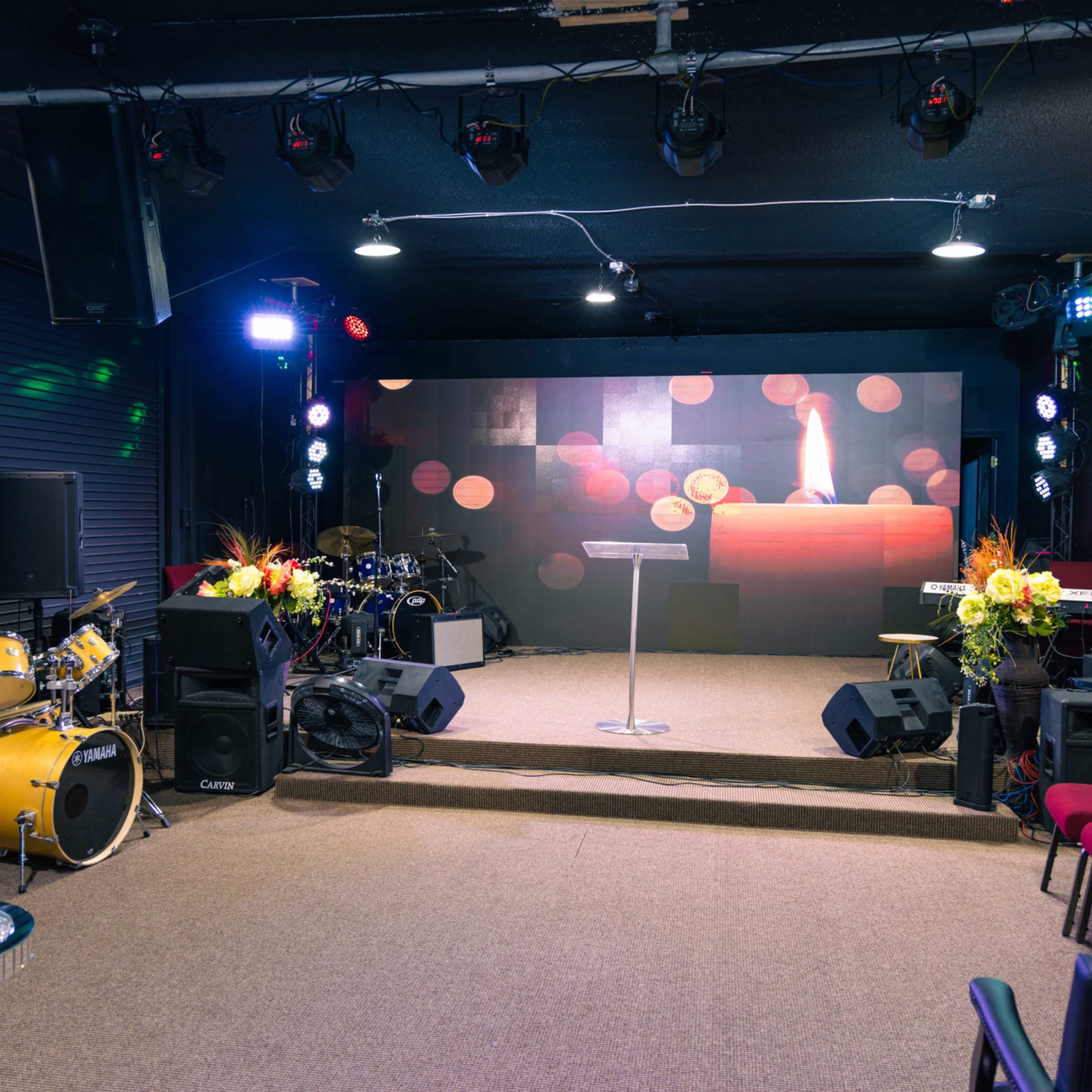 A small stage with a podium and a candle backdrop, surrounded by musical instruments and chairs arranged in front.