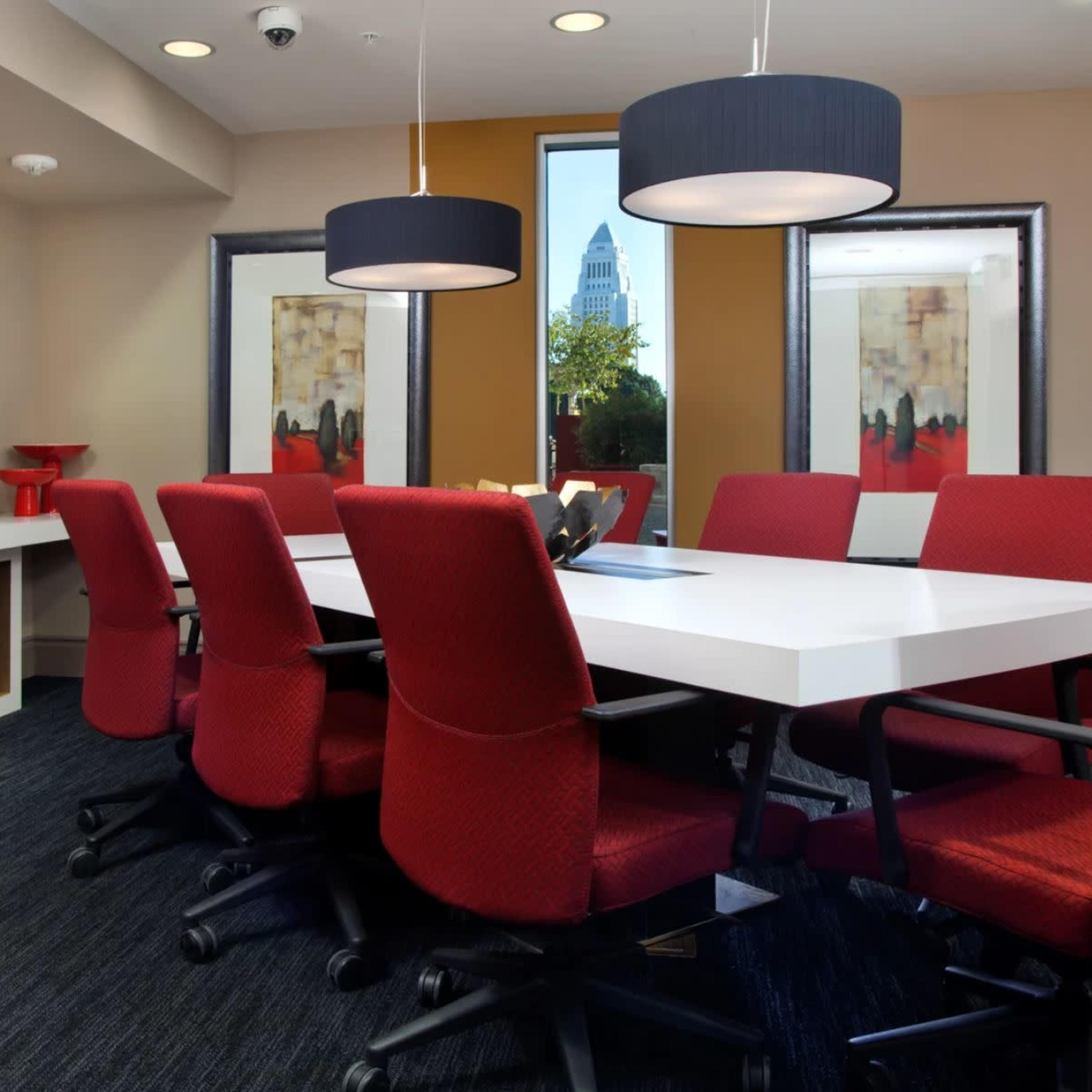 A modern conference room features a large white table surrounded by red chairs, with a TV on the wall and windows showing a city view.