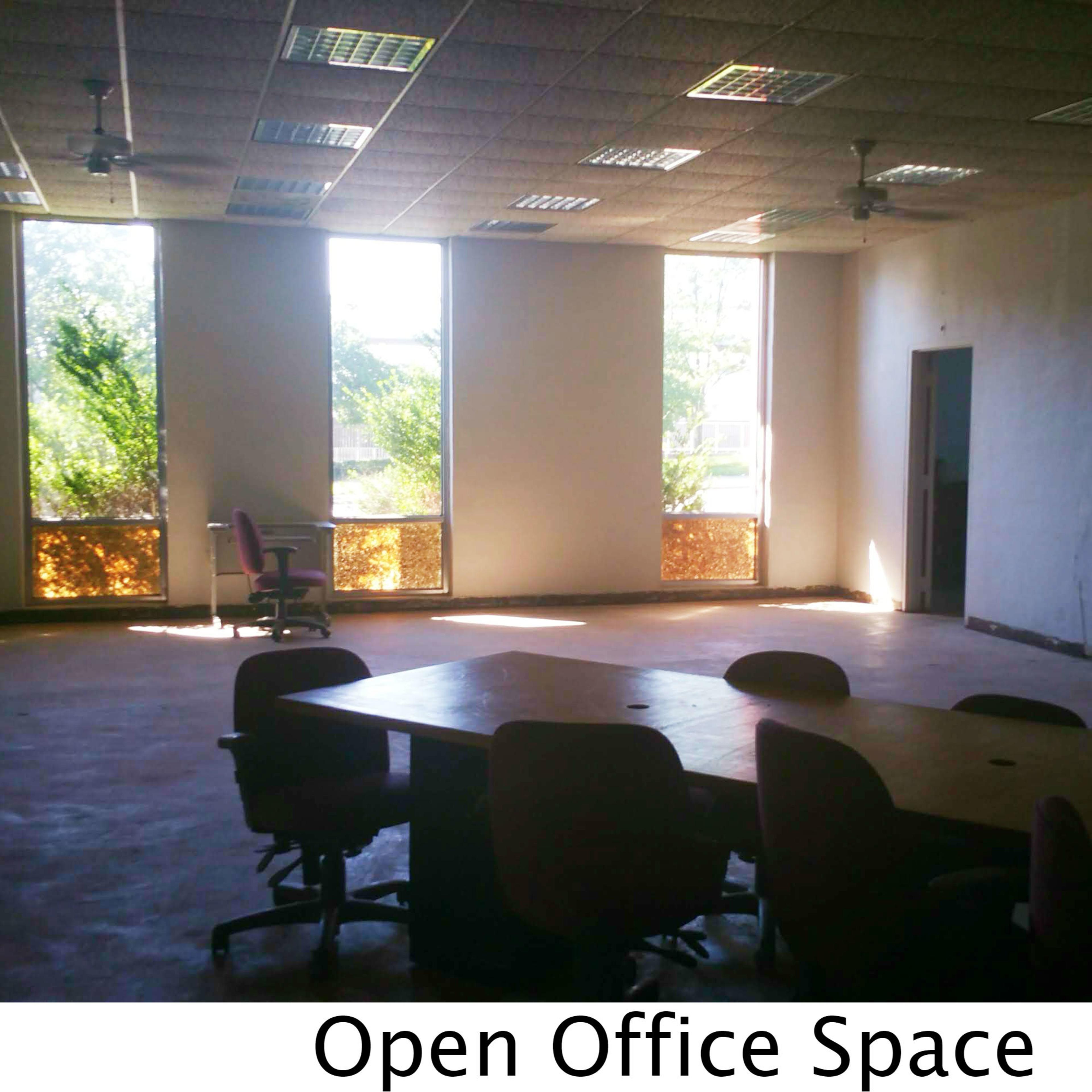 An open office space featuring a large table surrounded by several chairs, with large windows allowing natural light to enter.