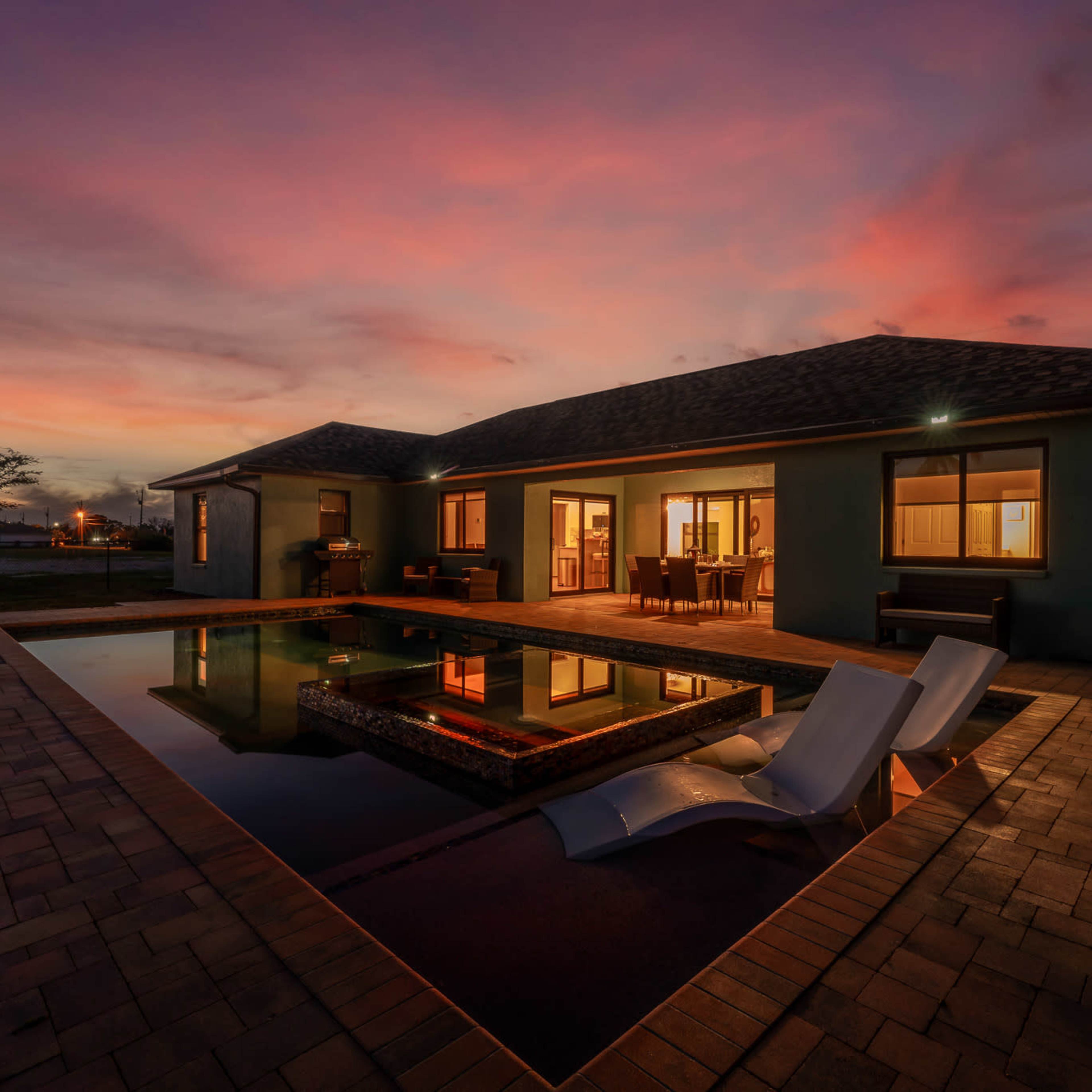 A modern house with a swimming pool reflects a colorful sunset sky in the background.