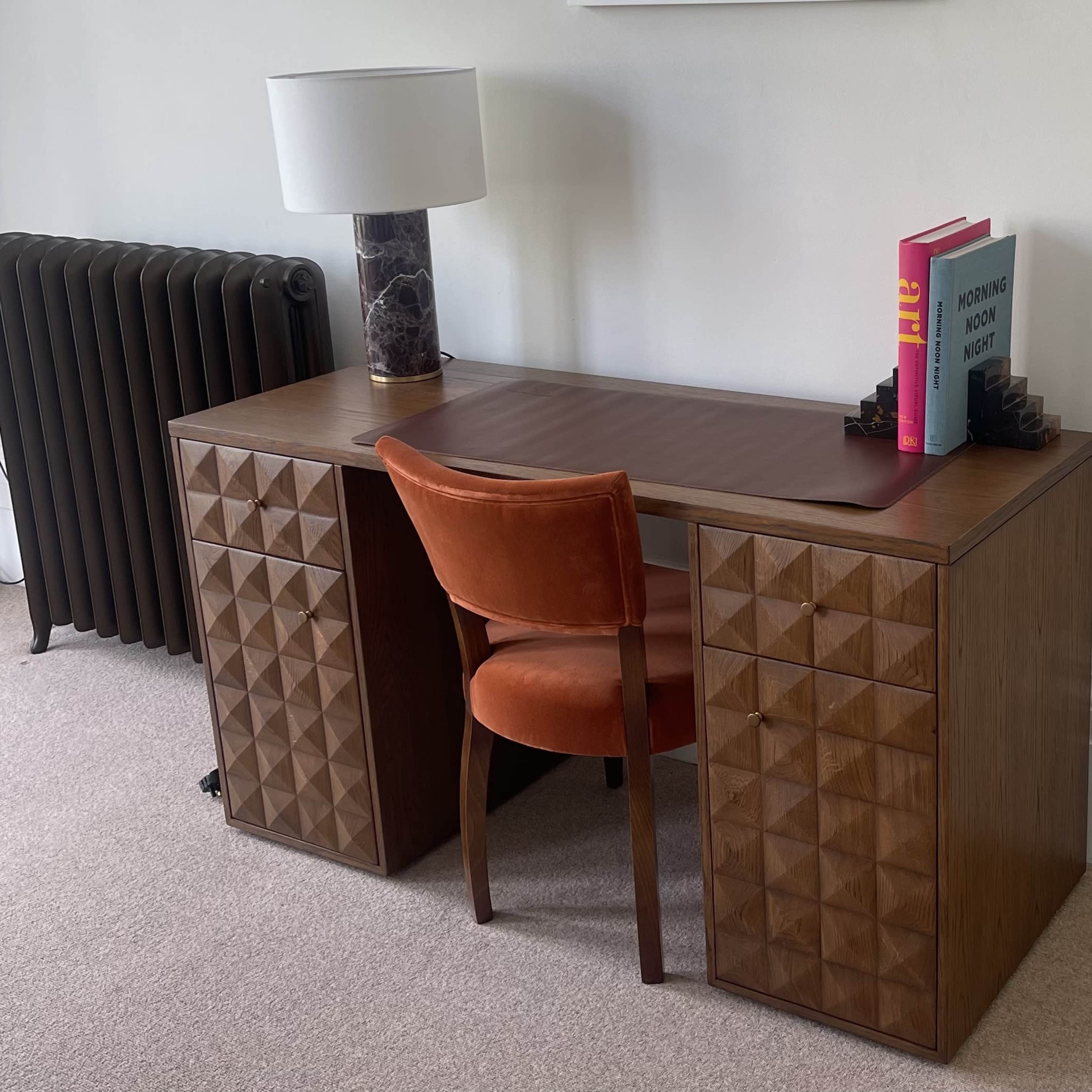 A wooden desk with a textured front, a round chair in orange upholstery, a table lamp, and books on the desktop, set against a neutral wall and a radiator.