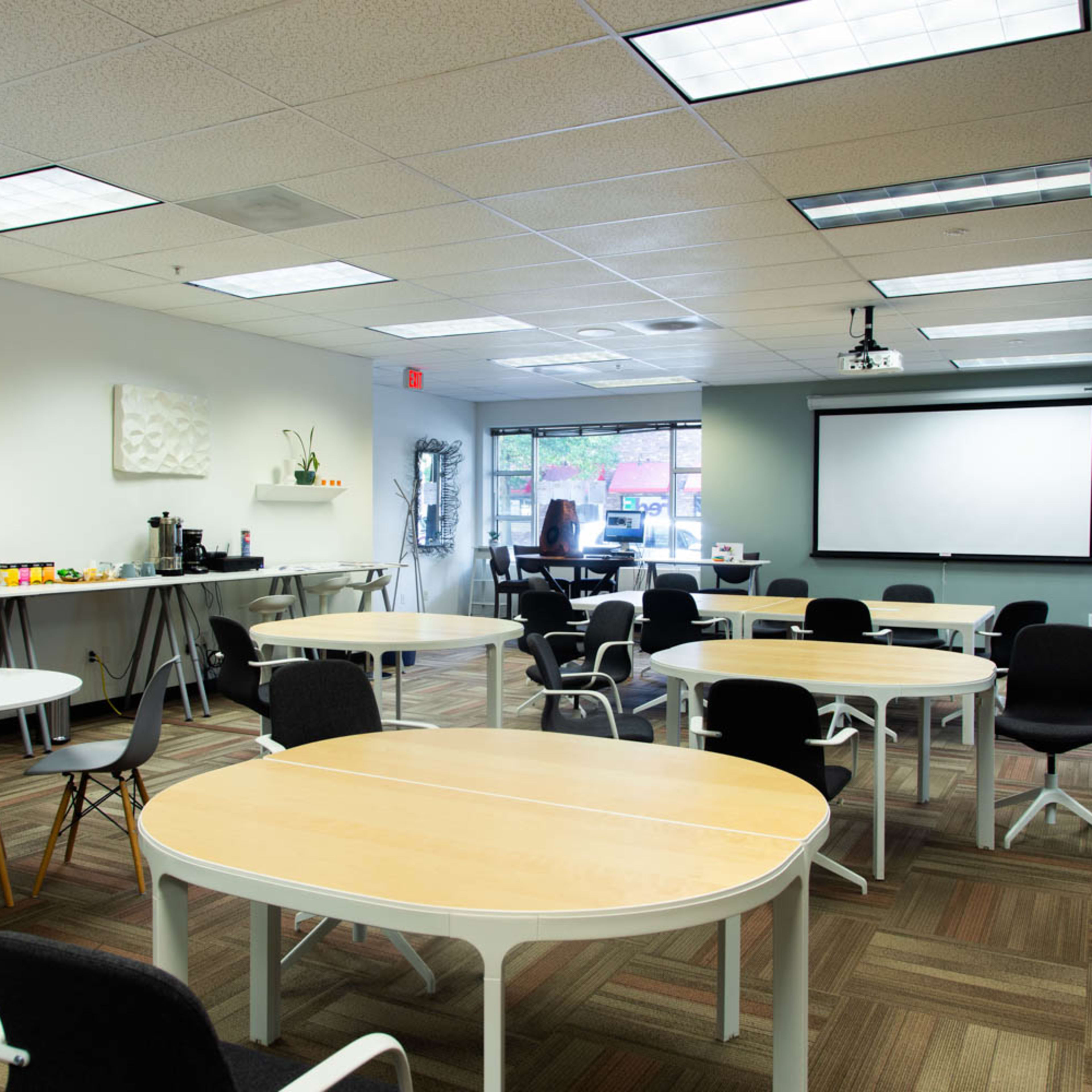 A modern office conference room with several round and rectangular tables arranged, a projector on the ceiling, and a whiteboard at the front.
