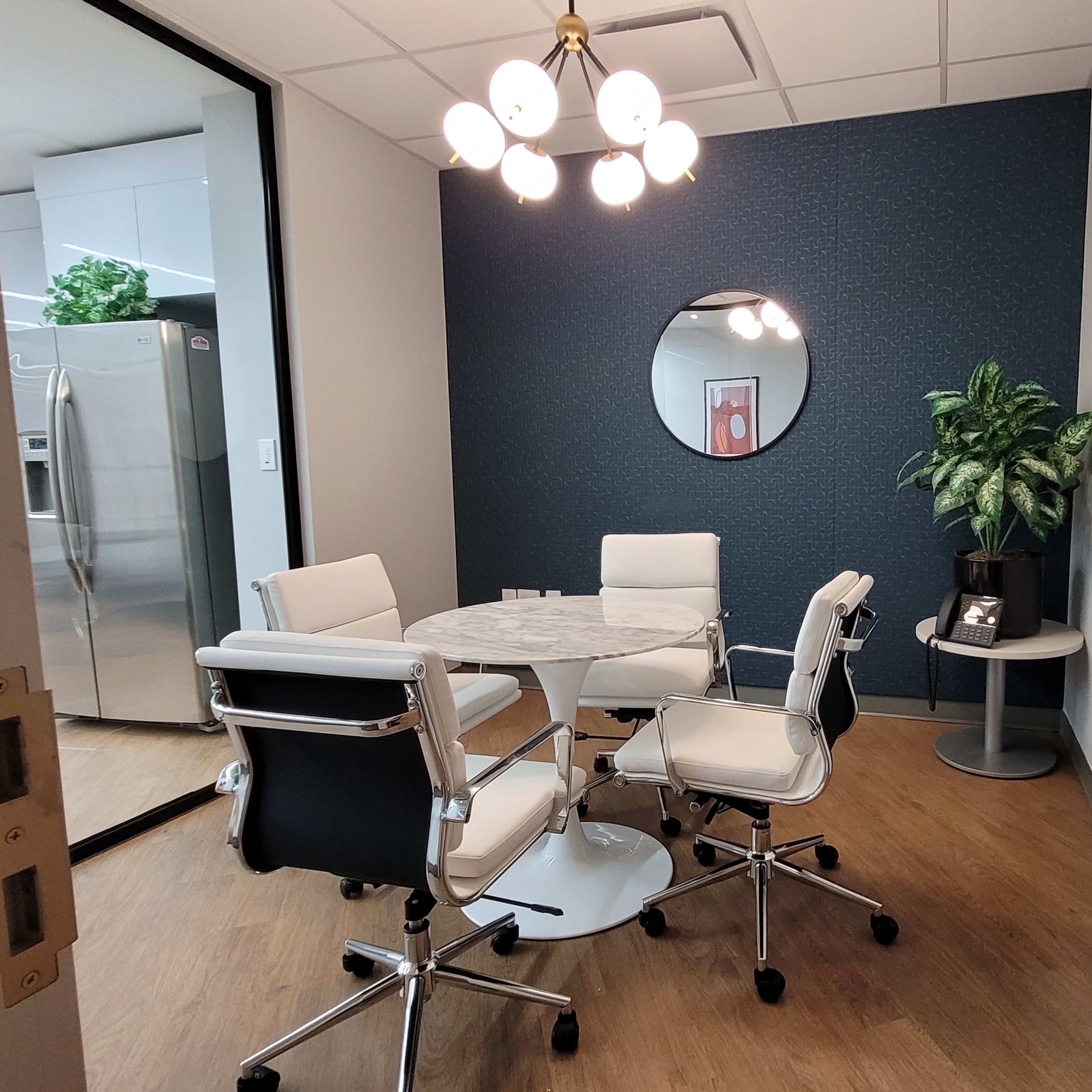 A small meeting room with a round table, four white chairs, a potted plant, and a large mirror on the wall.