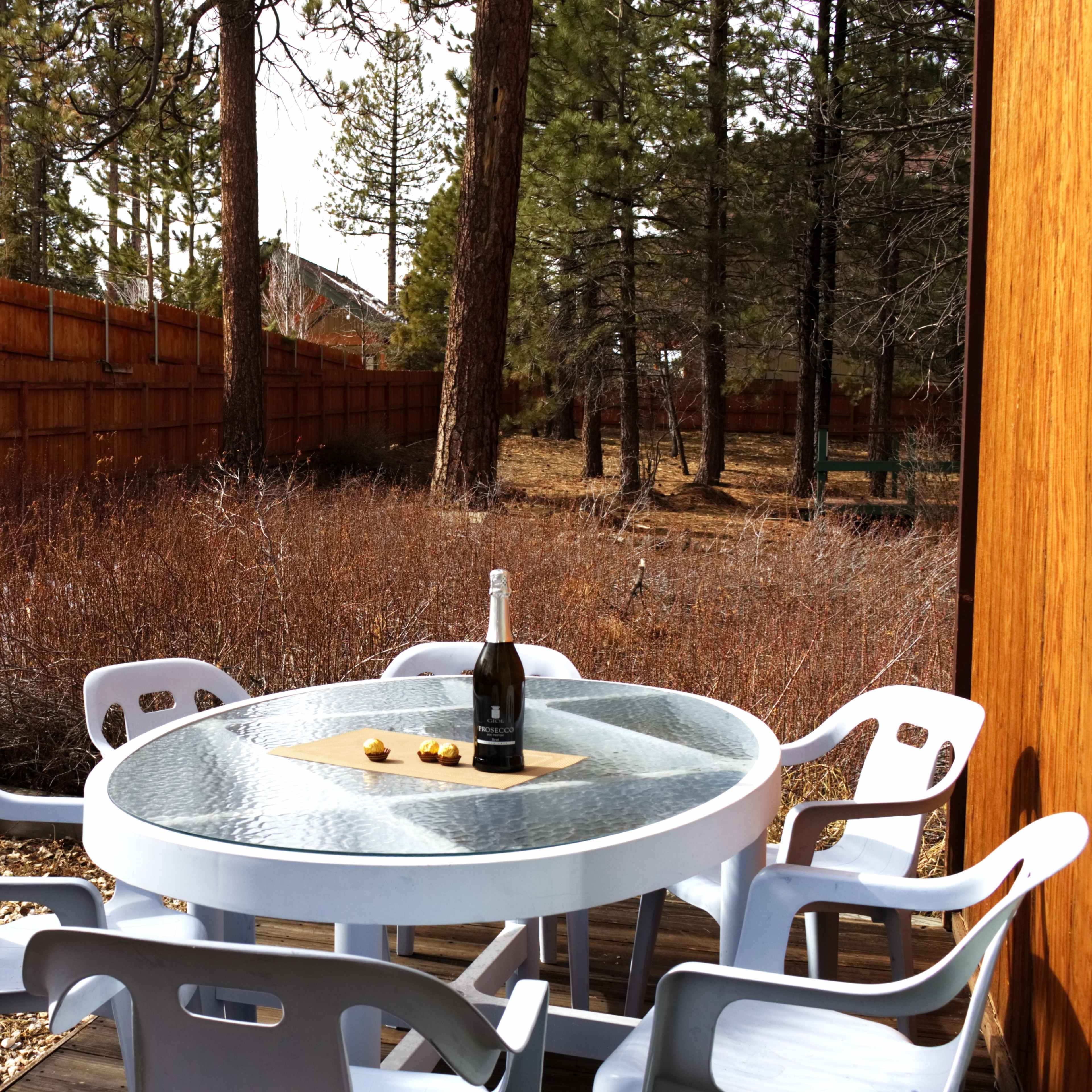 A glass-top table with a bottle of champagne and a small plate sits surrounded by white plastic chairs on a wooden deck in a wooded backyard.