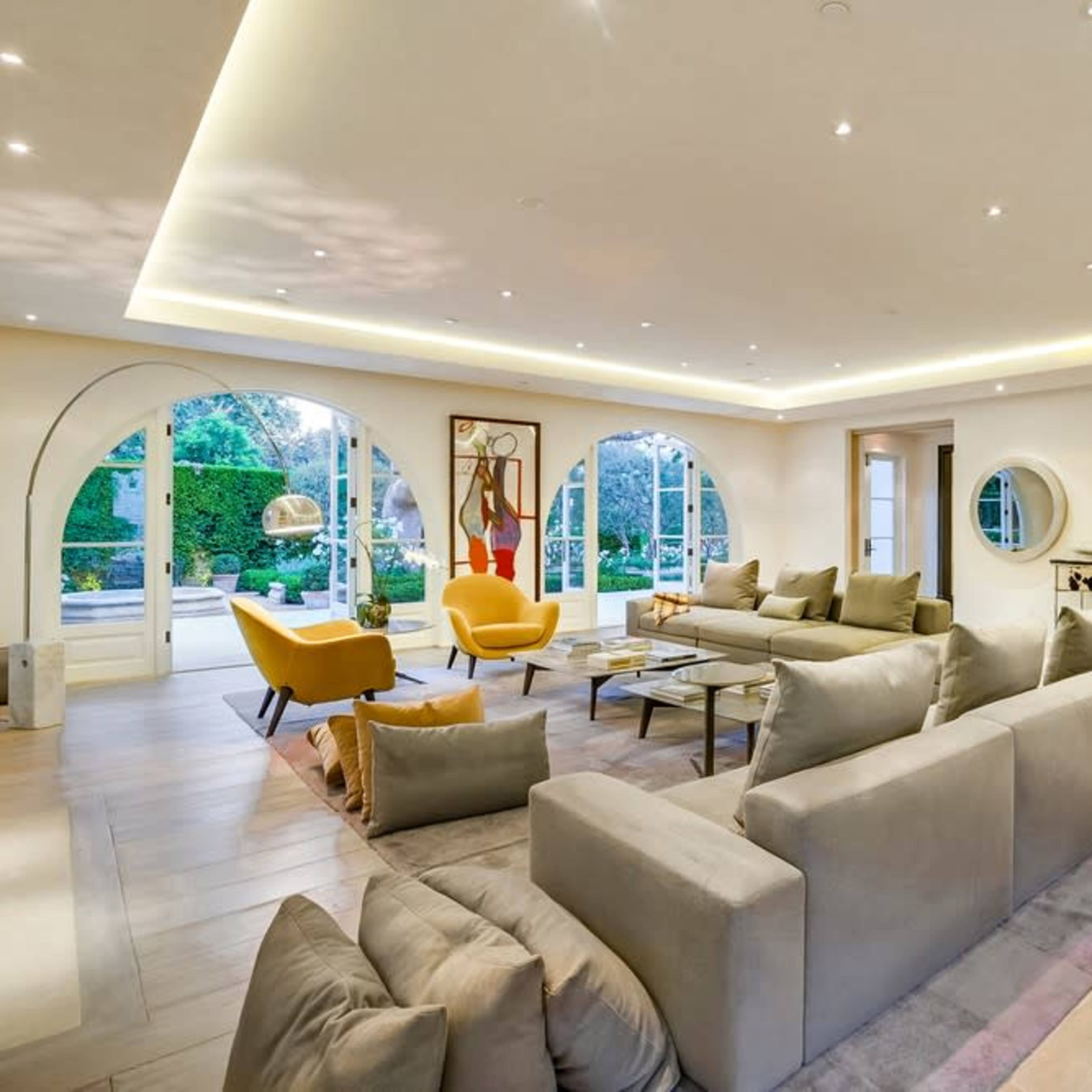 A modern living room with a large sectional sofa, yellow armchairs, and a sleek kitchen in the background, illuminated by recessed lighting and large windows that allow natural light.