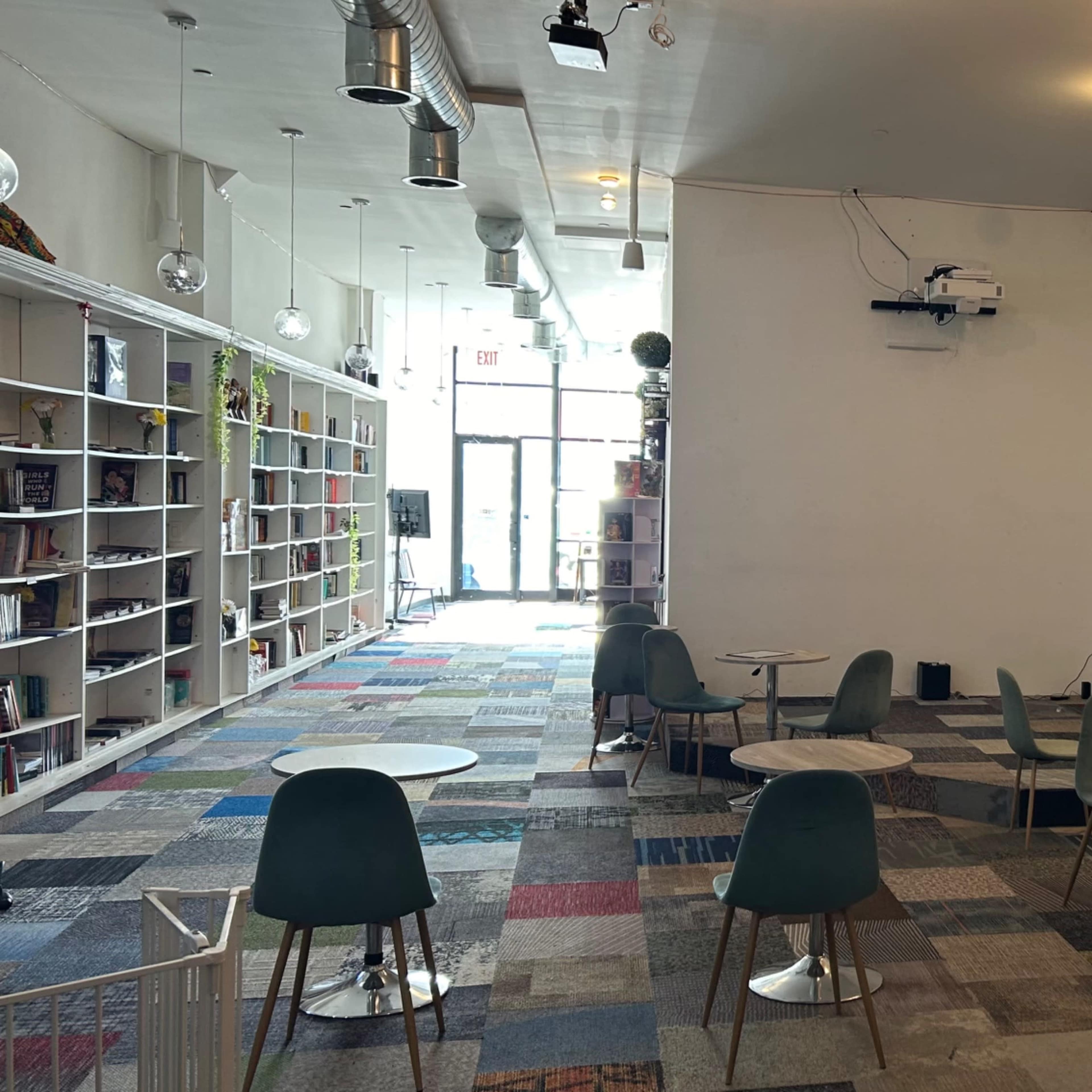 A spacious cafe area featuring several small round tables and chairs, with a large blank screen on one wall and shelves filled with books.
