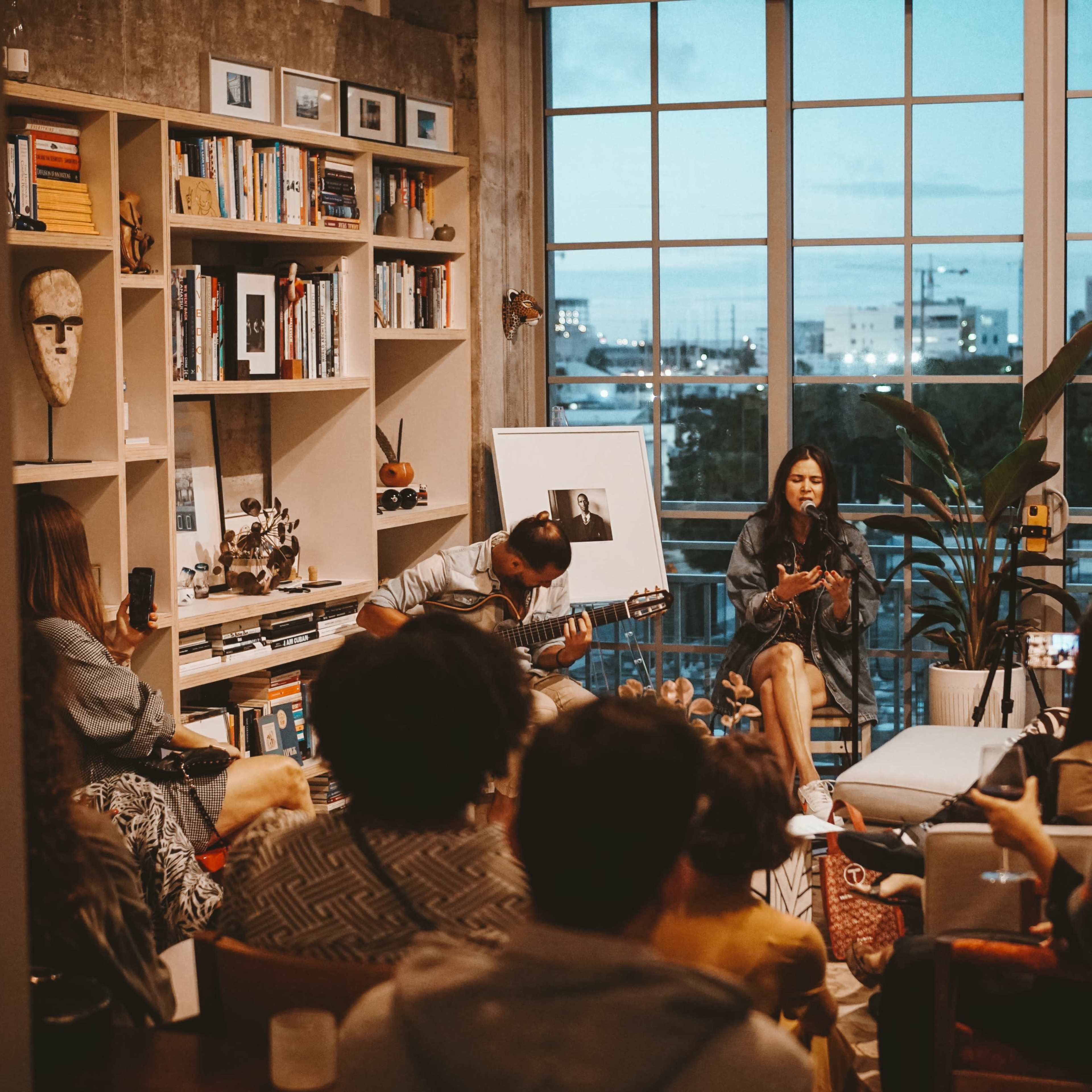 A woman sings and plays guitar while seated in a cozy living room filled with people listening and enjoying the performance, with a large window showcasing a city skyline at dusk.
