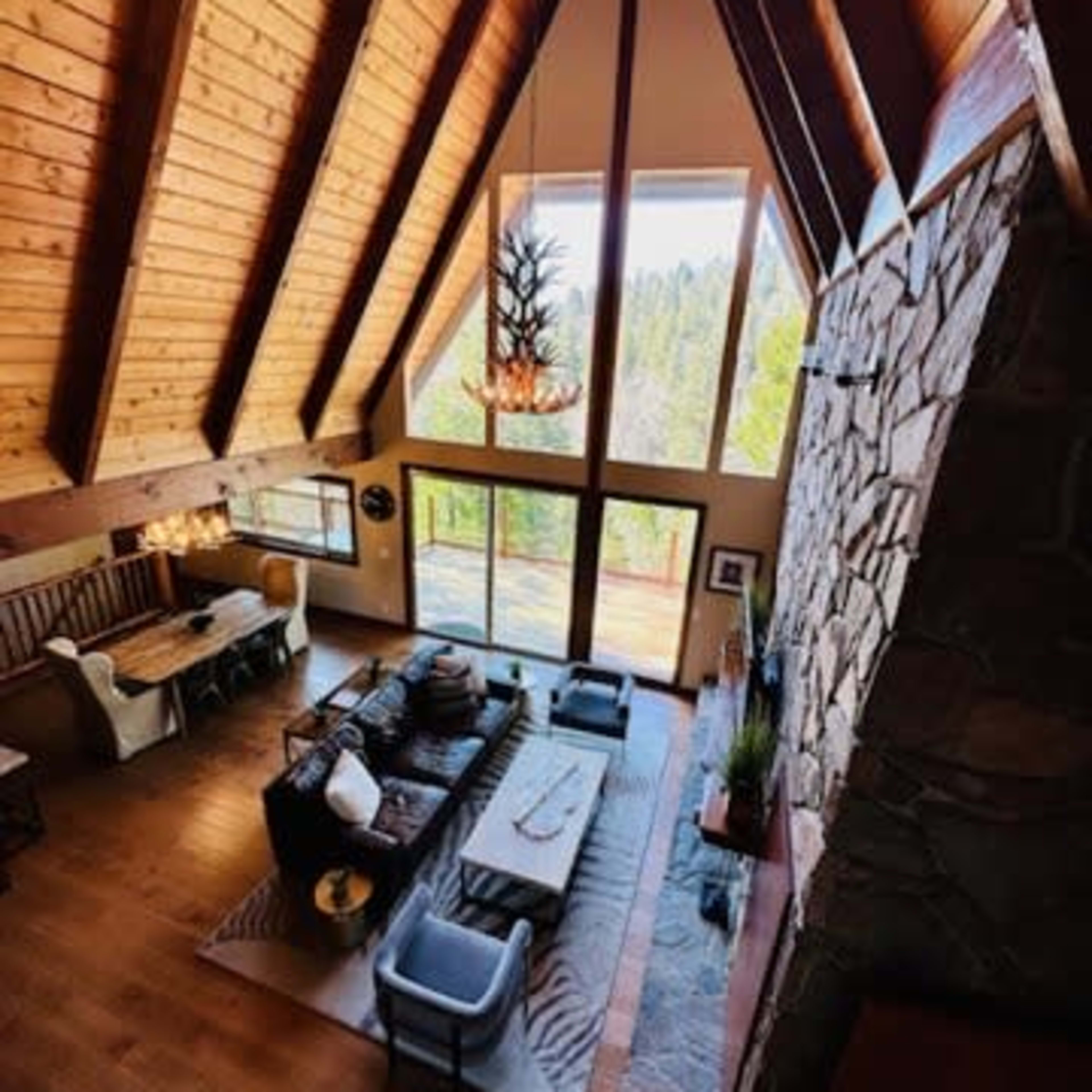 A spacious, open-concept living area features a high, wooden A-frame ceiling, large windows, and a stone fireplace, complemented by contemporary furniture.