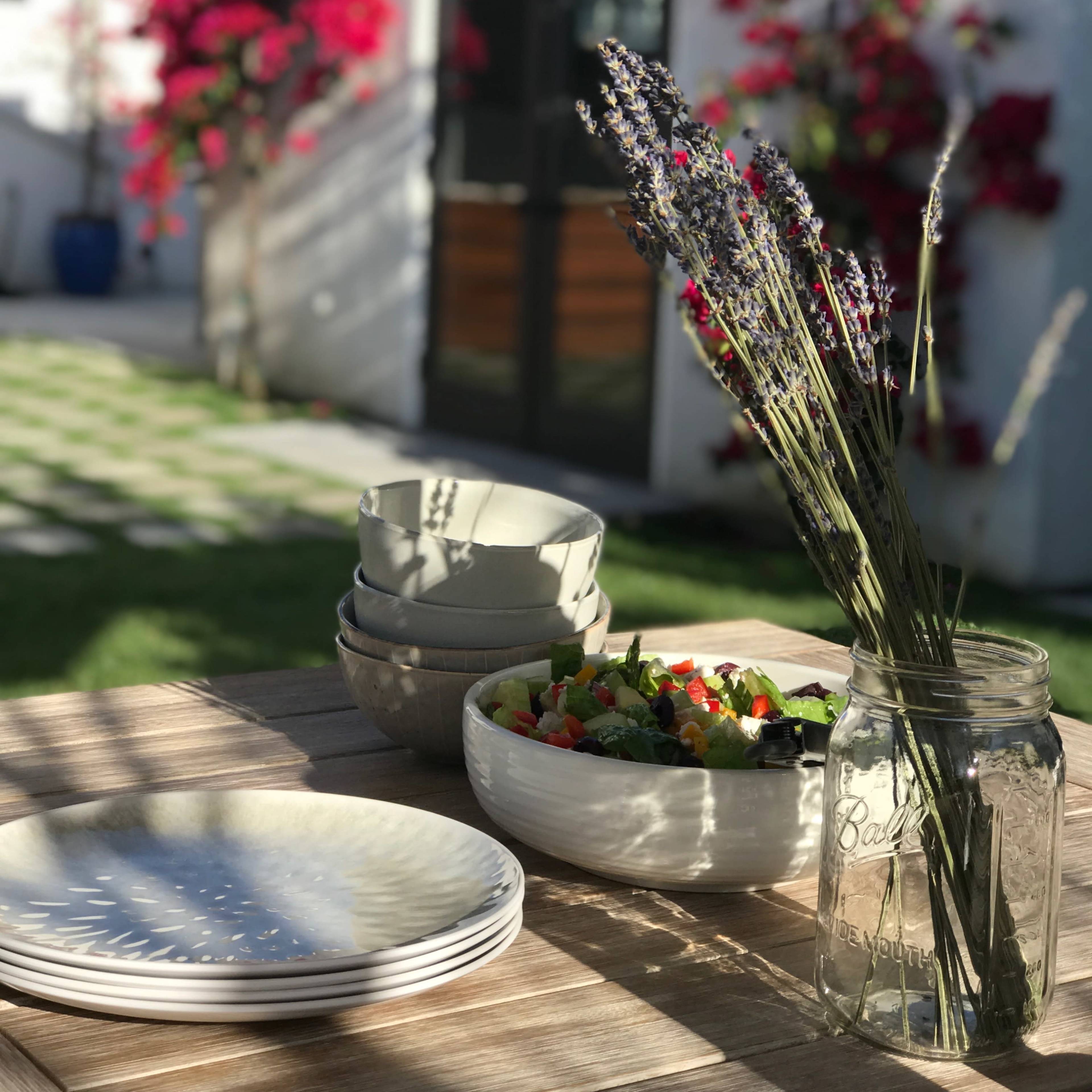 A wooden table is set outdoors with bowls and a salad, alongside a vase of lavender, while vibrant flowers decorate the background.