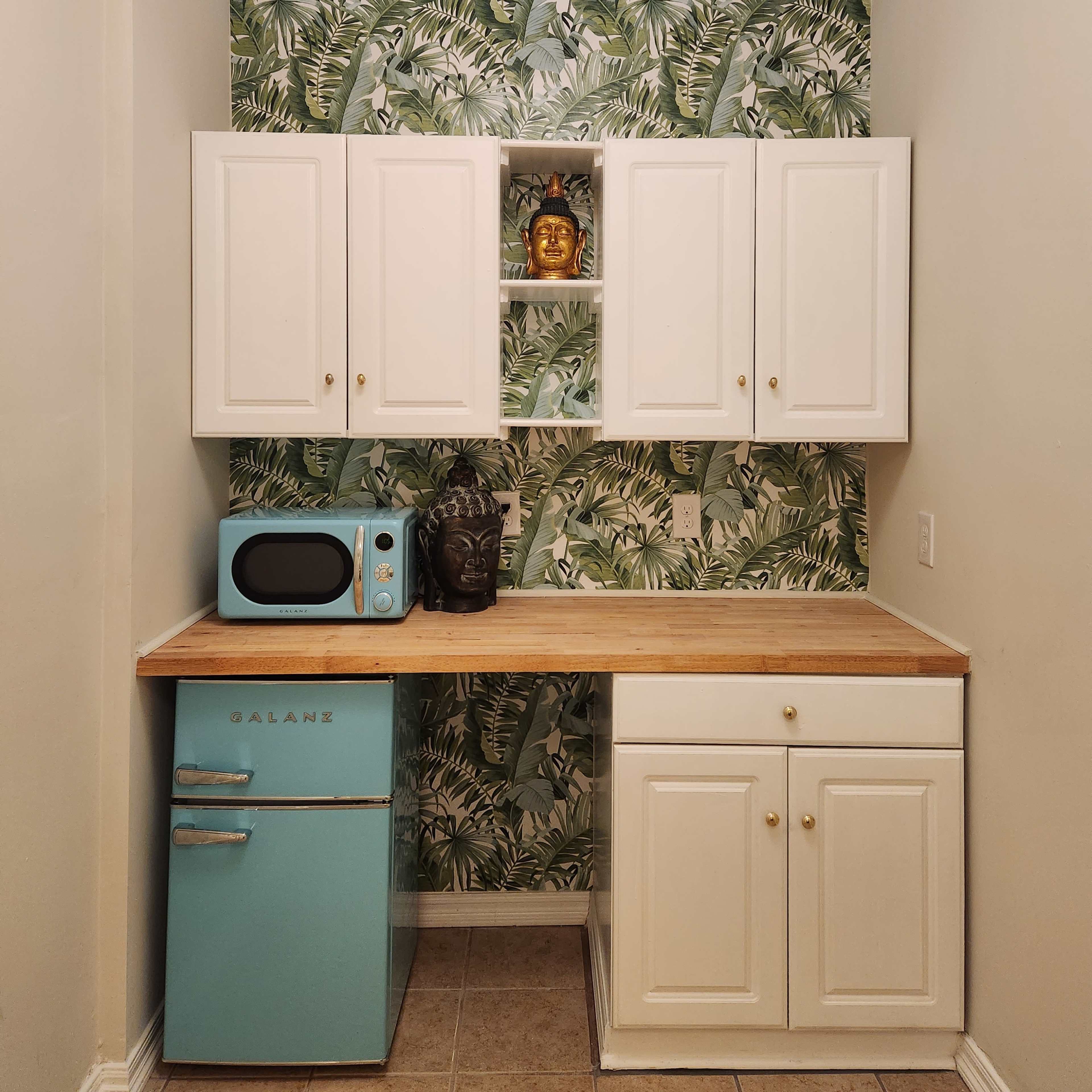 A small kitchen area featuring a light blue fridge, white cabinets, a microwave, and a patterned green wallpaper with palm leaves.