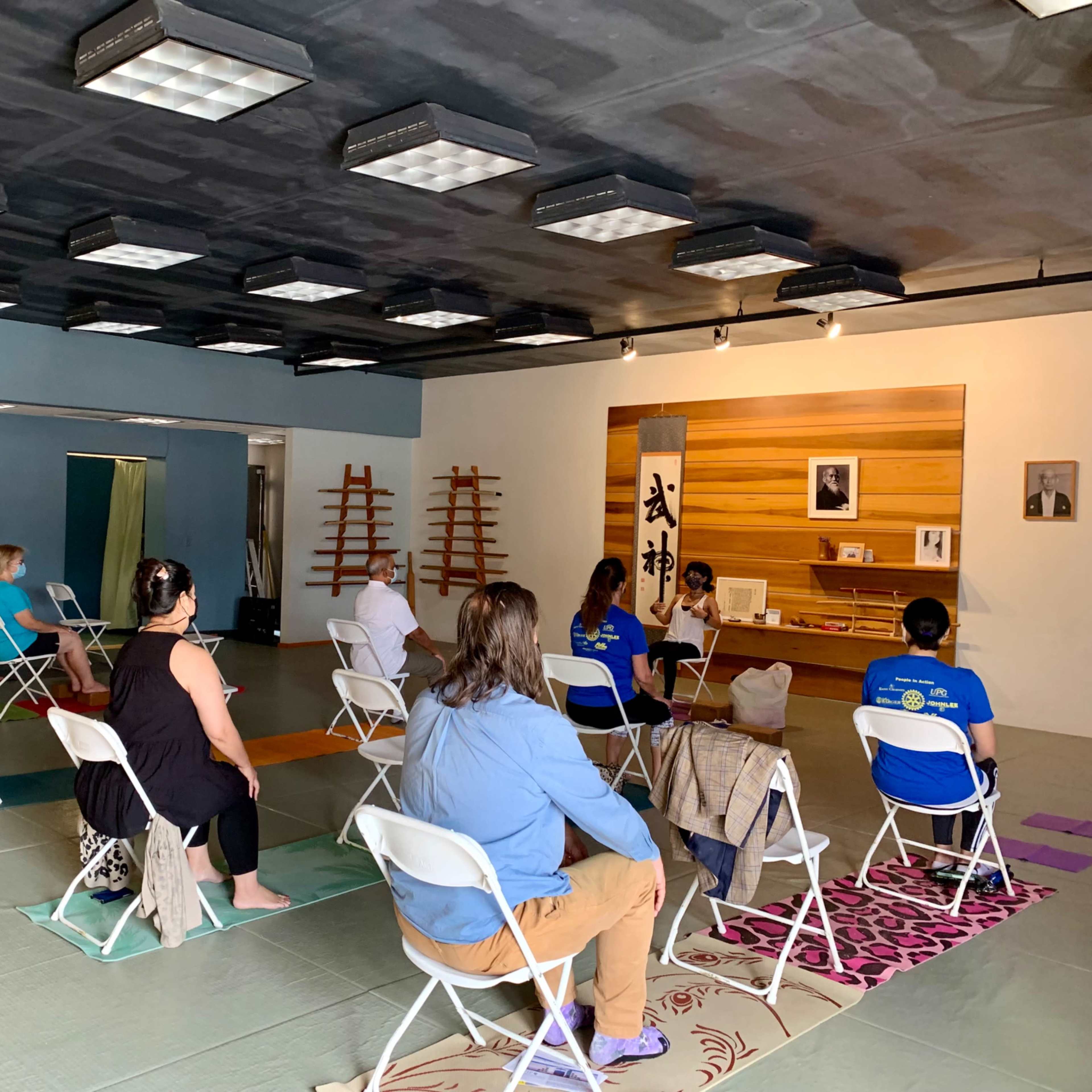 A group of people sits on folding chairs in a spacious room with tatami mats, facing a small altar and a wall displaying an image and text.
