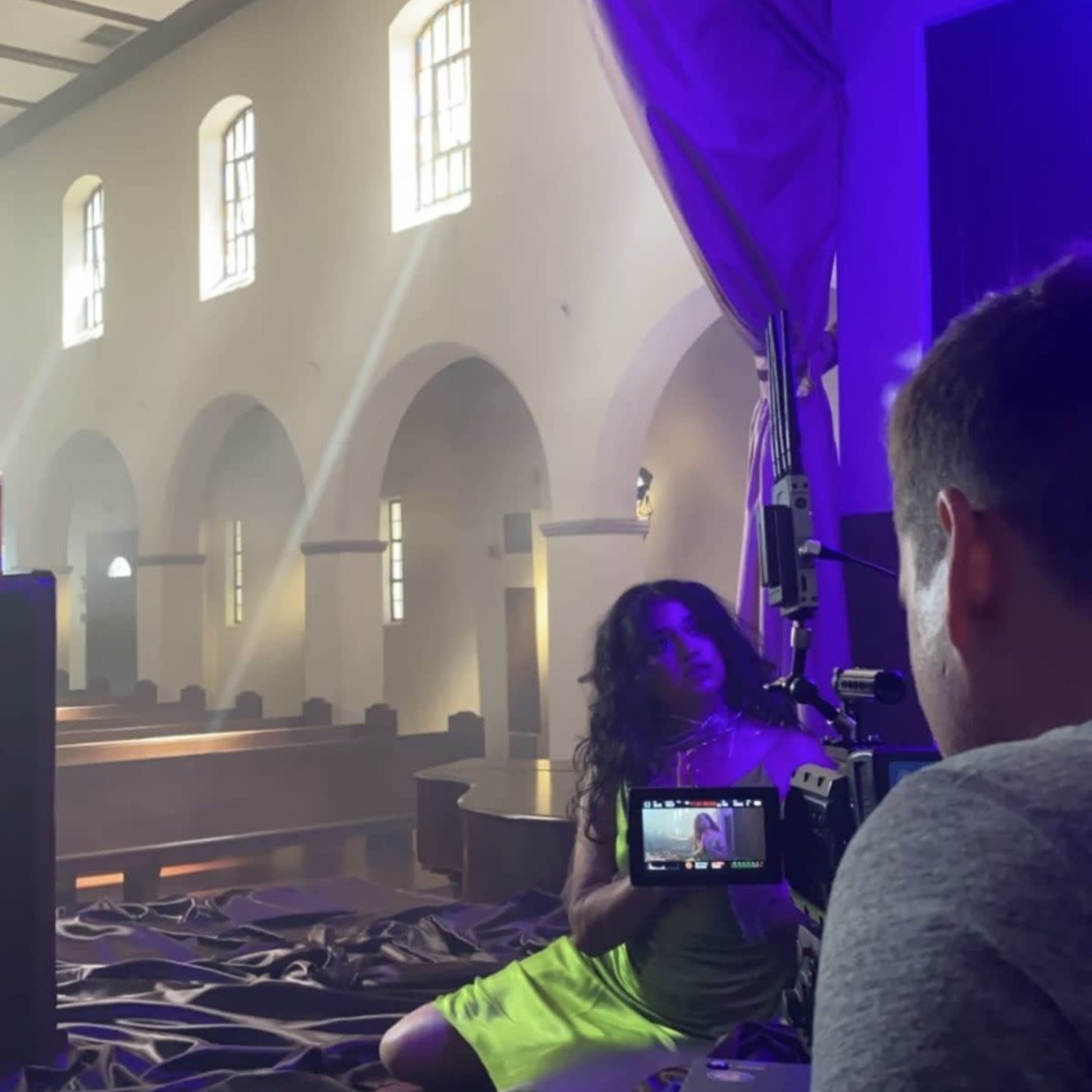 A woman in a bright yellow outfit sits on the floor of a church while a man records her with a camera.