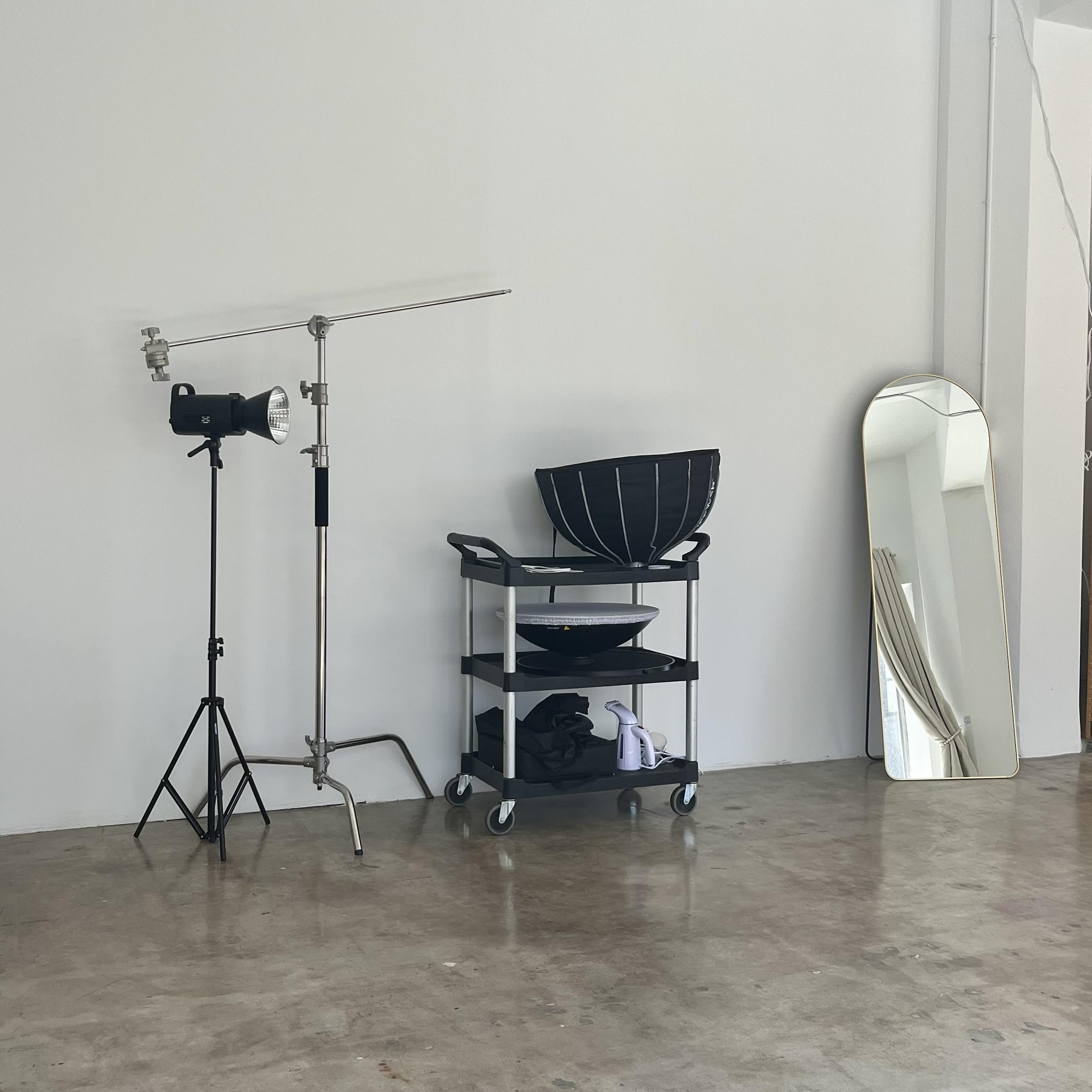 A studio corner with a light stand, a cart holding two bowls, and a tall mirror against a plain wall.