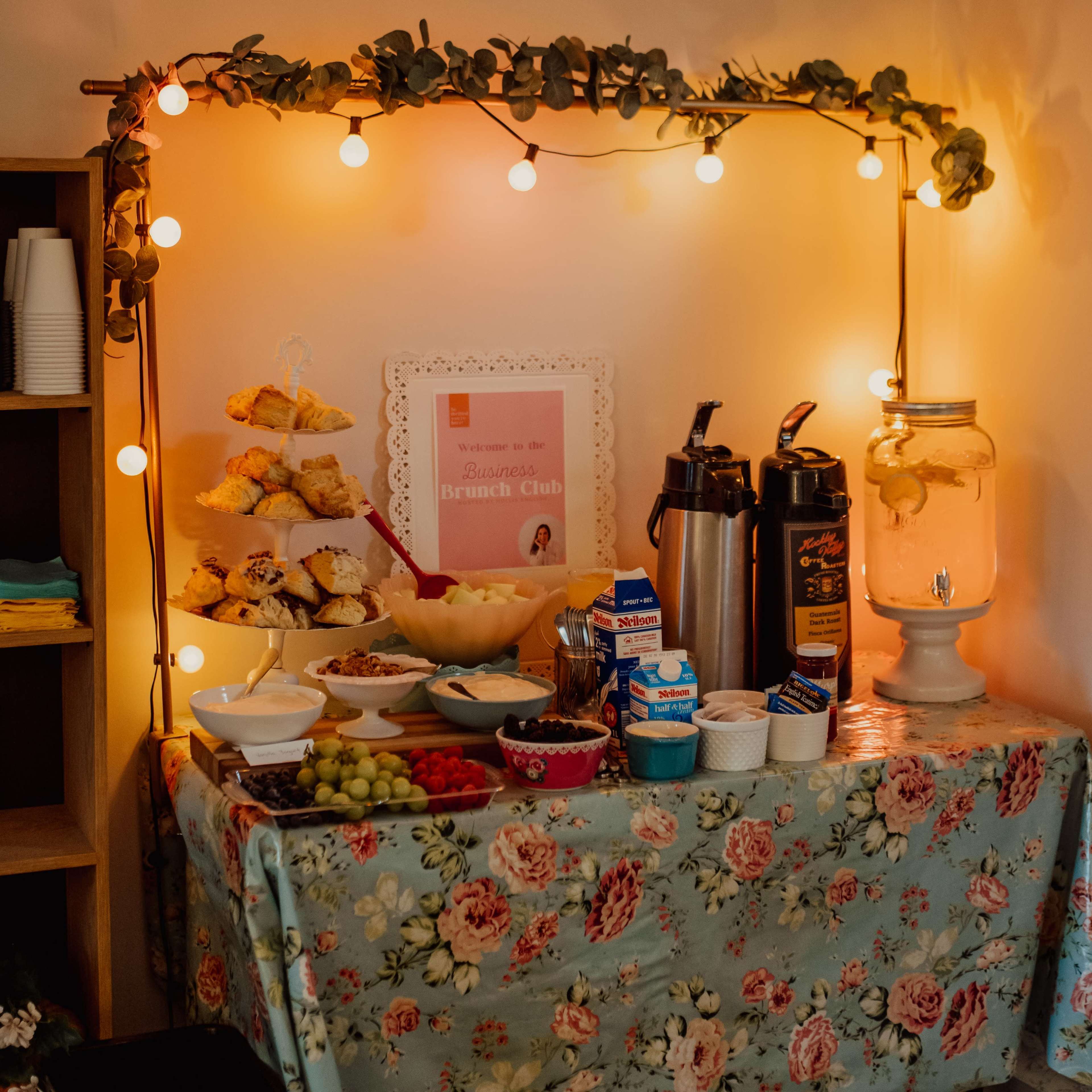 A table is set up with food and drinks, including pastries, fruit, and several beverage containers, adorned with string lights and colorful tableware.