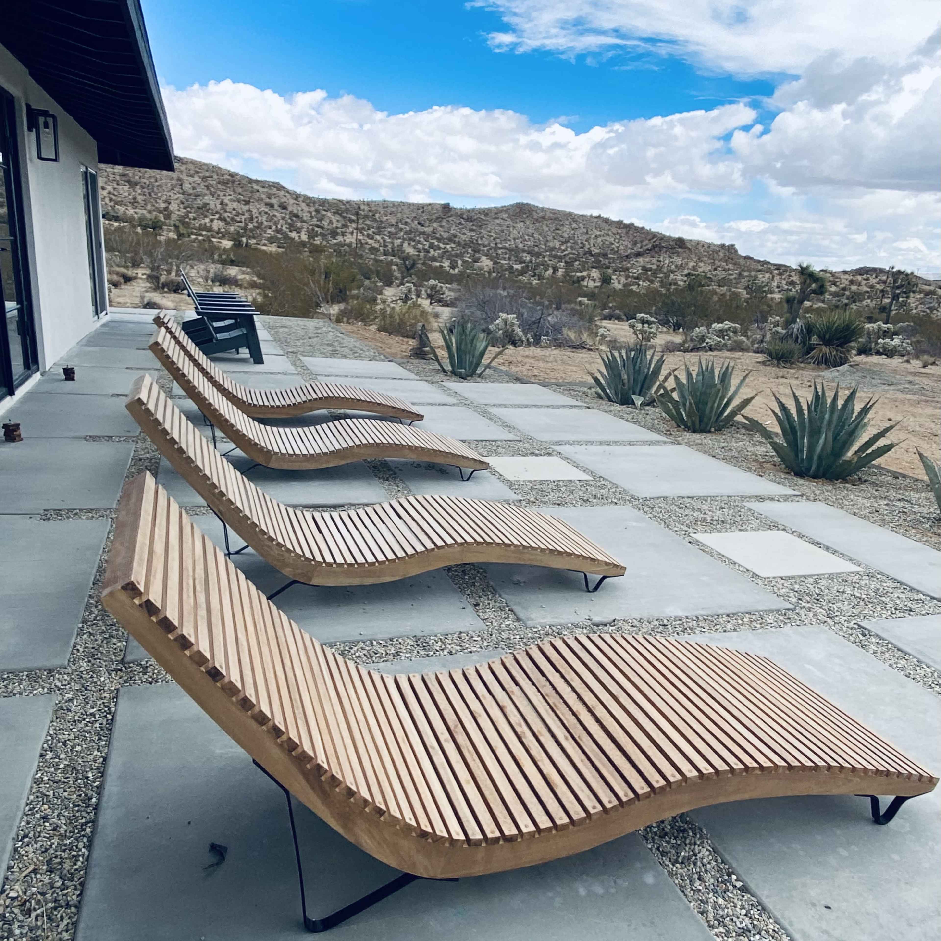 Four wooden lounge chairs are arranged on a stone patio overlooking a desert landscape with sparse vegetation and rolling hills under a cloudy sky.