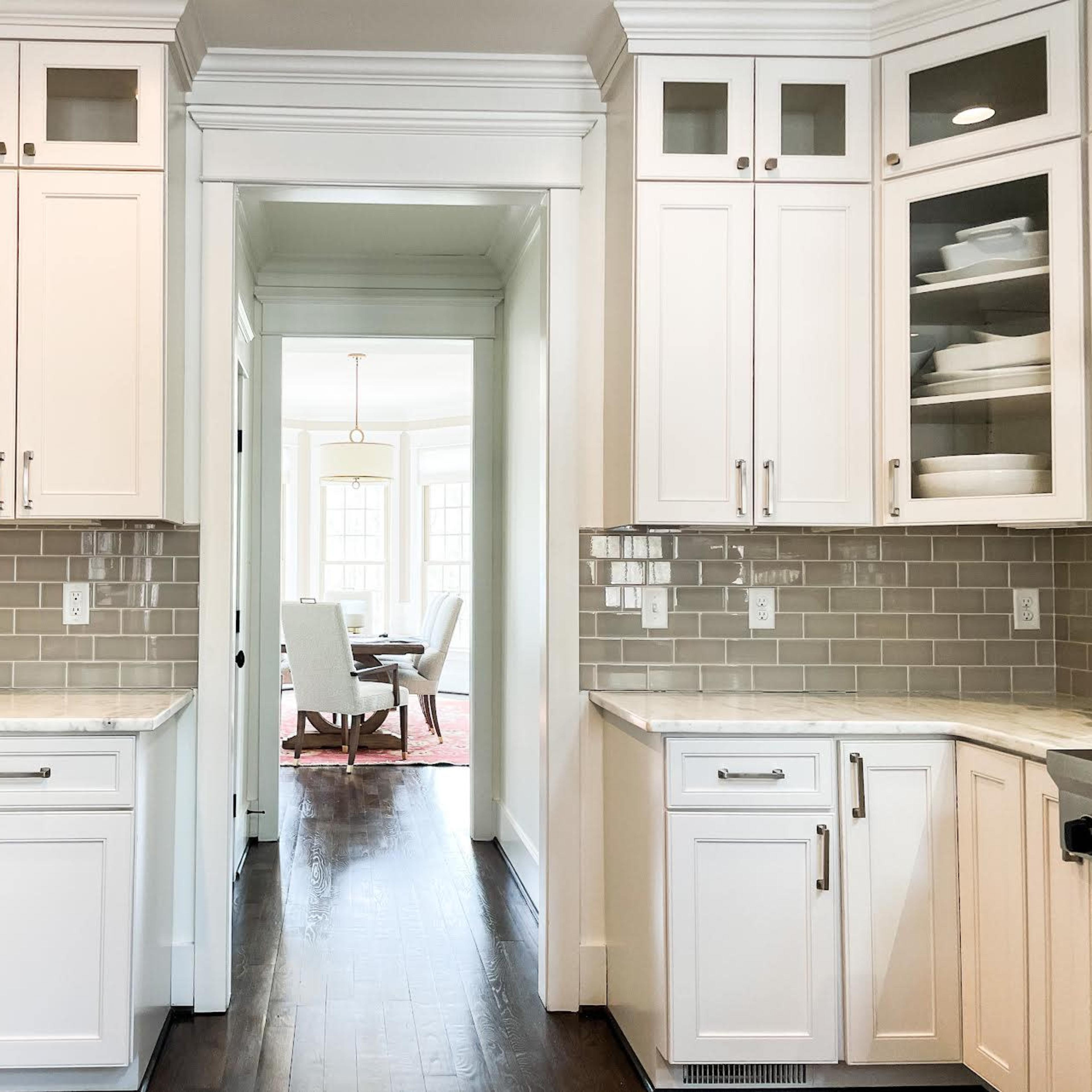 A bright kitchen featuring white cabinets and a light gray tiled backsplash, leading into a dining area visible through an open doorway.