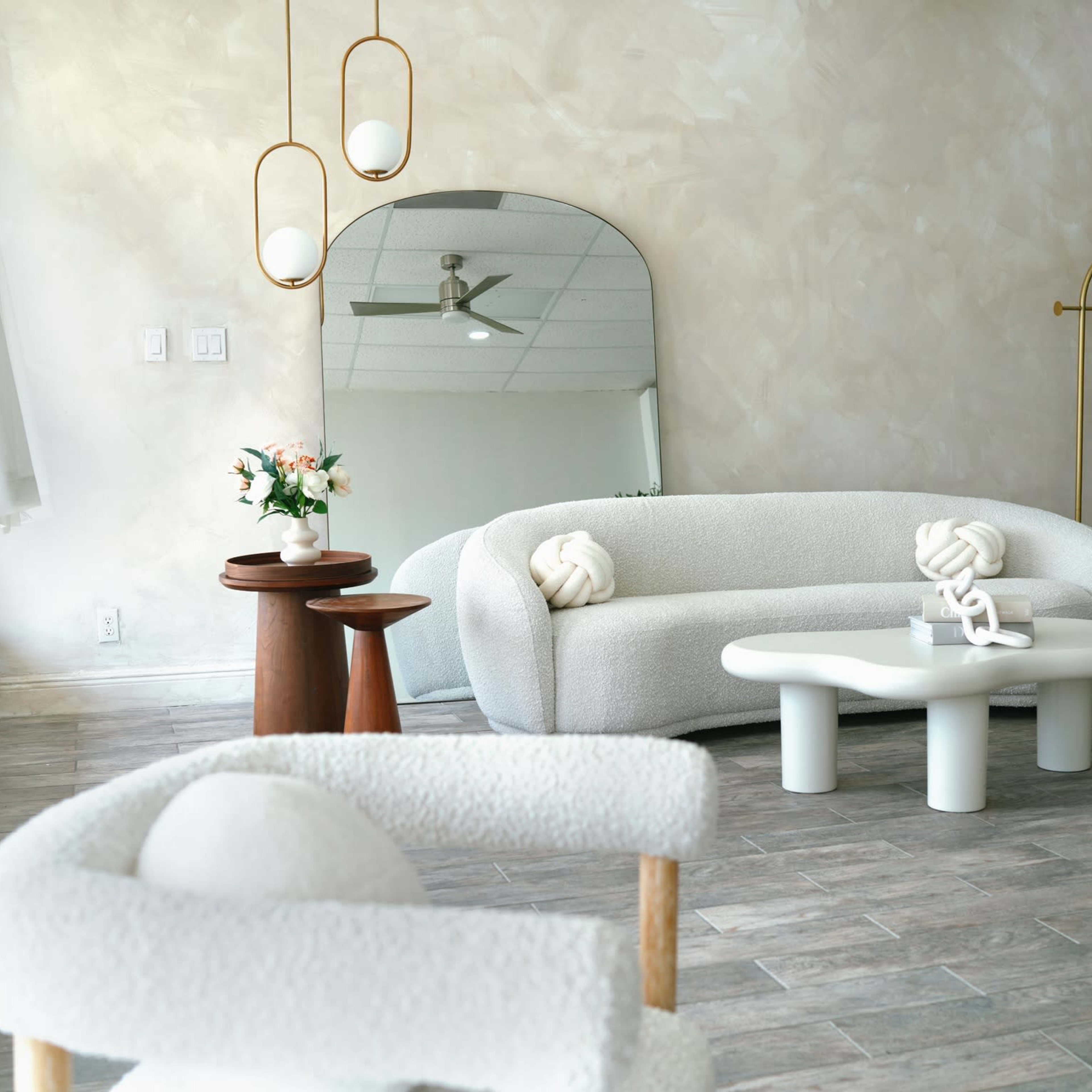 A modern interior space featuring a curved white sofa, a round wooden table, a low white coffee table, and decorative elements like flowers and mirrors against a softly colored wall.