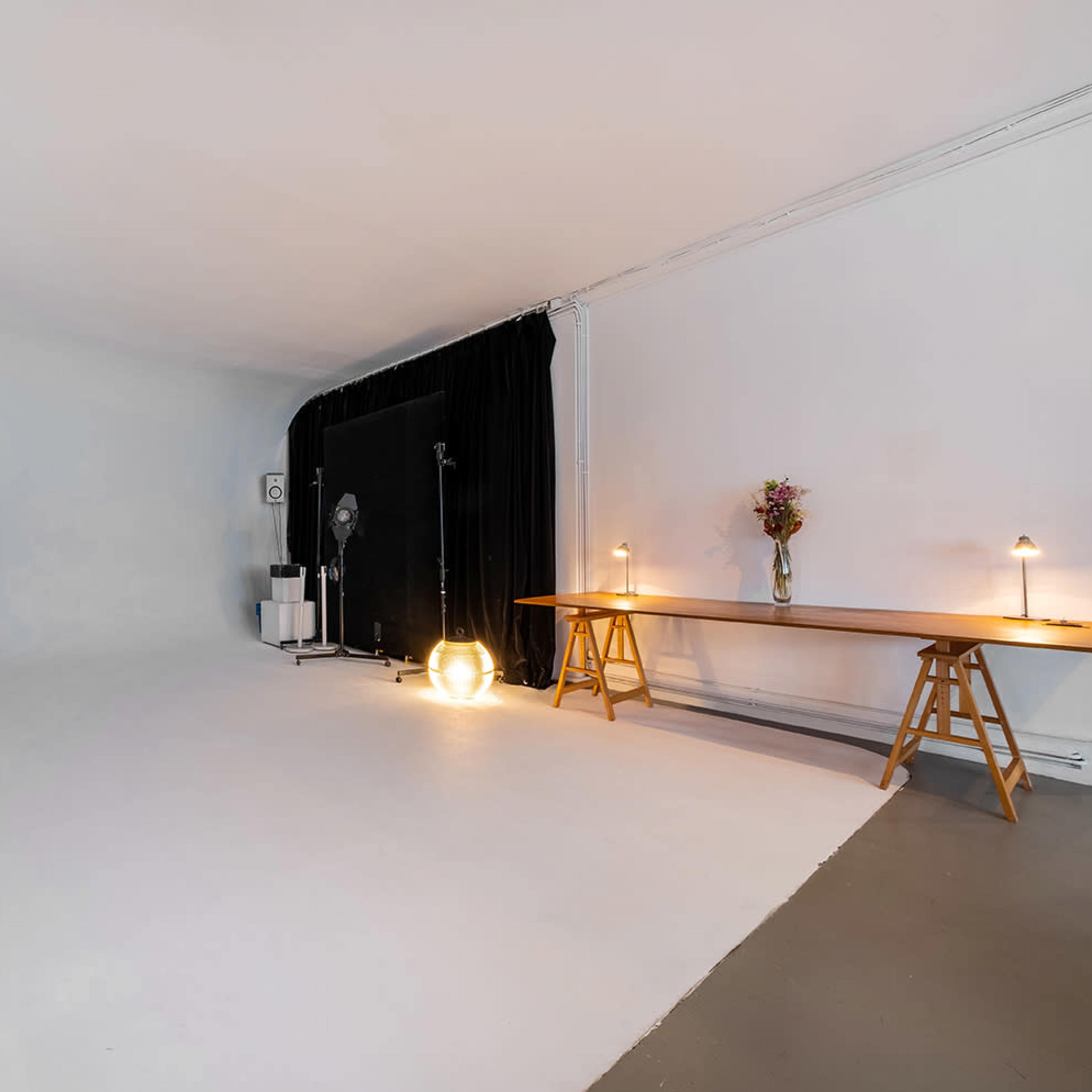 A spacious, minimalist studio with a plain white backdrop, black curtains on one side, and a wooden table with lamps and a vase of flowers.