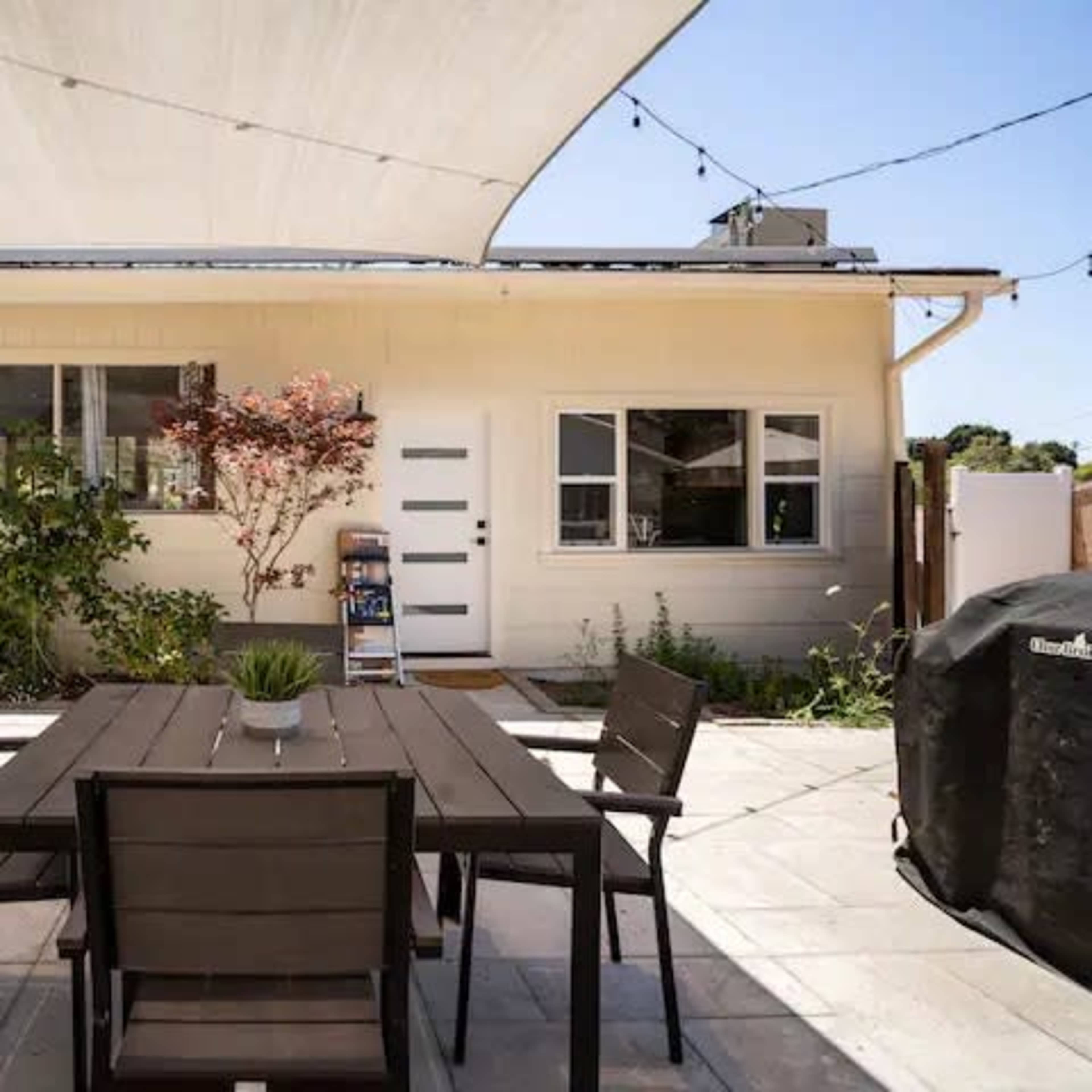 A patio area featuring a wooden table with chairs, surrounded by plants and a house with a shaded wall.