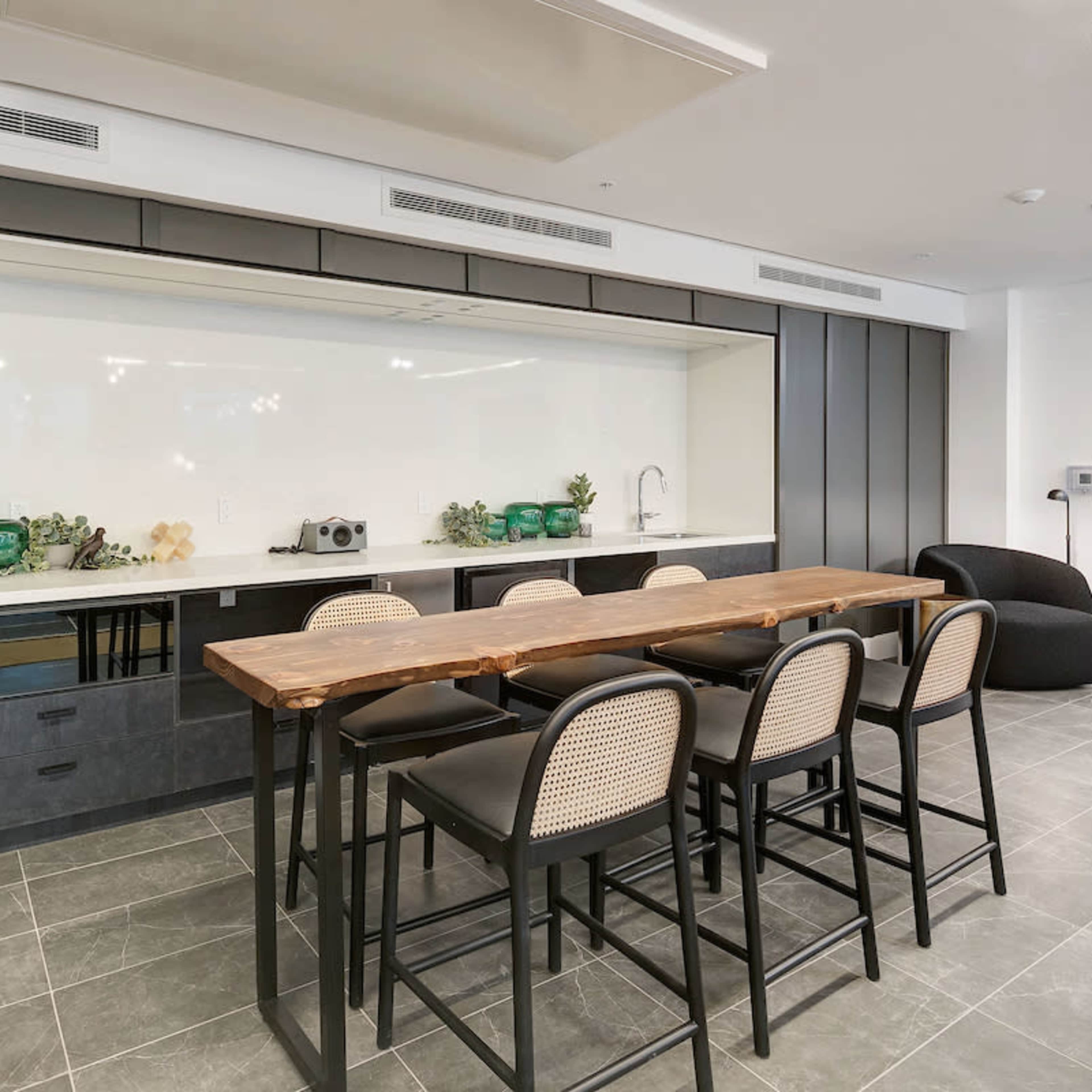 A modern kitchen features a long wooden table with black chairs, set against a sleek backsplash and minimalist cabinetry.