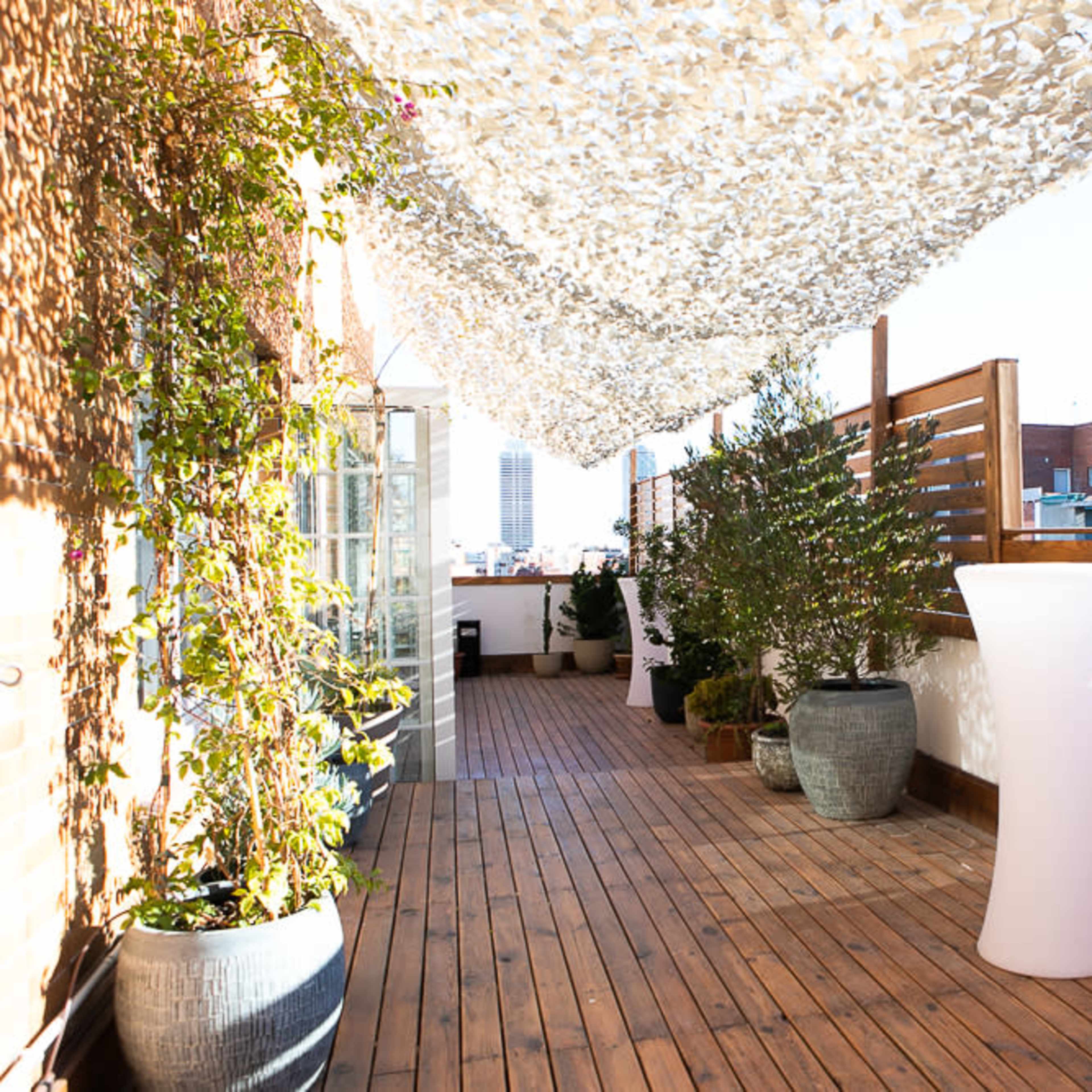A wooden terrace with potted plants, a white bar table, and a canopy overhead, overlooking a cityscape.