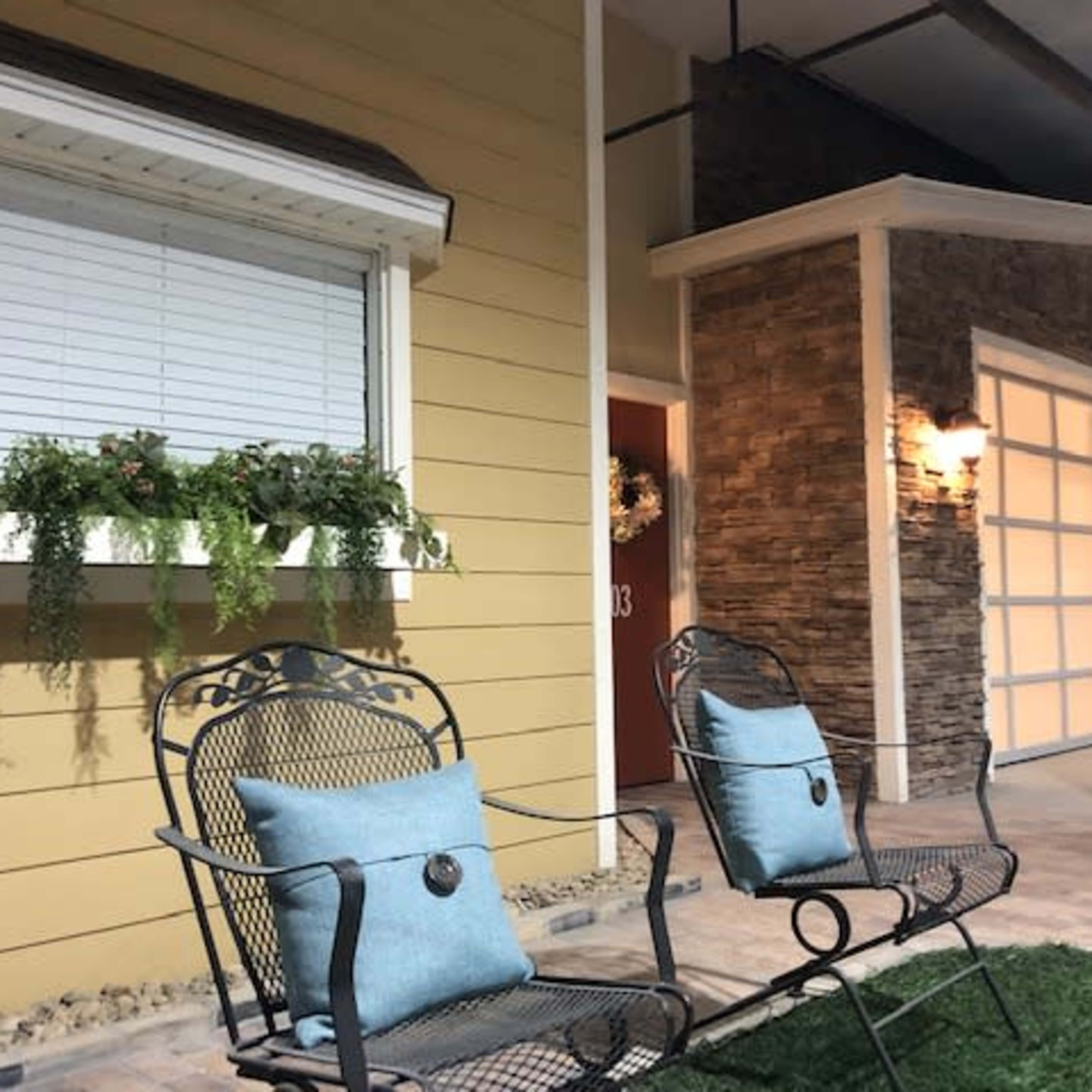 Two metal chairs with blue cushions sit on a small patch of grass in front of a house with a stone accent wall and a closed garage door.