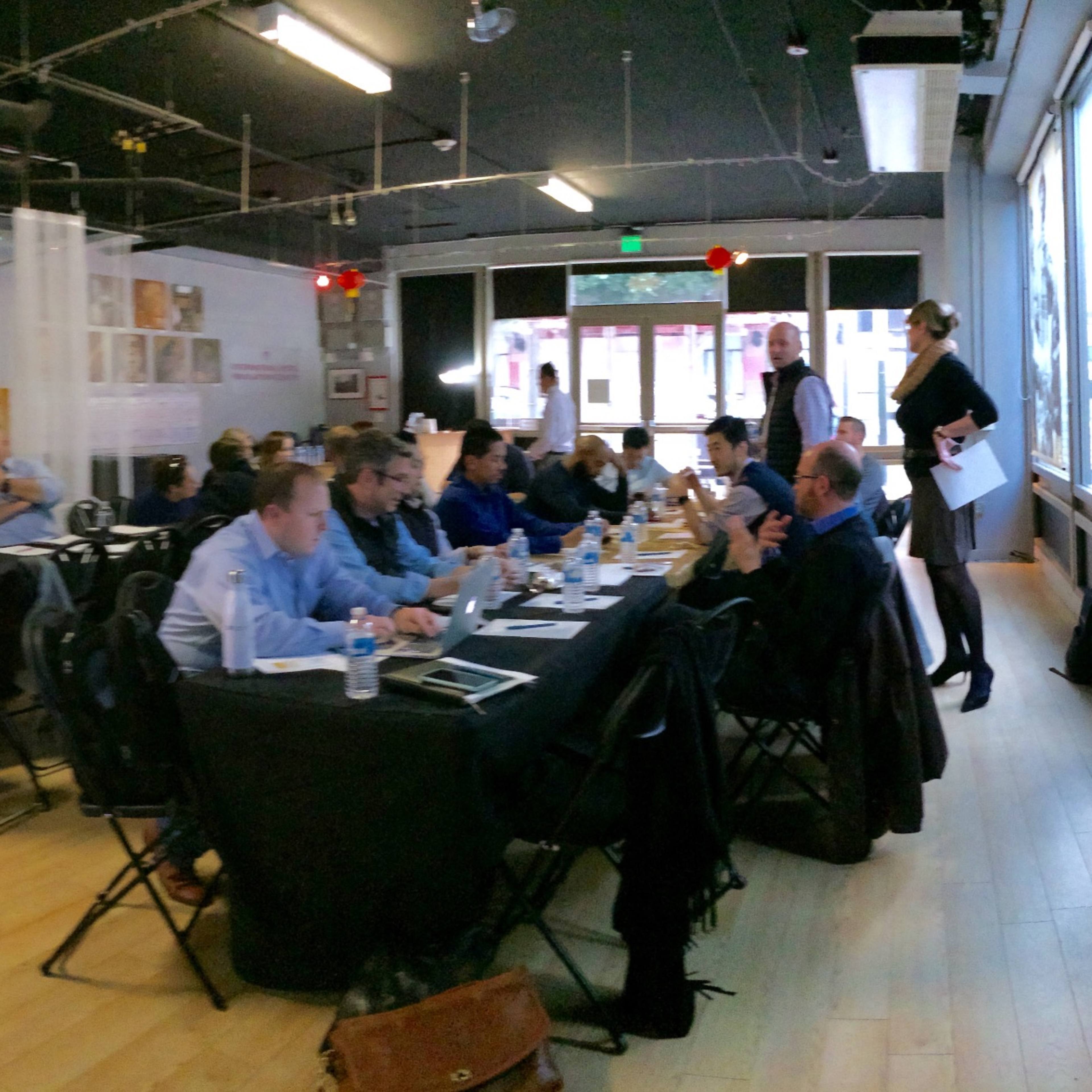 A group of individuals sits at a long table in a modern meeting space, with a projector screen displaying information and a whiteboard nearby.