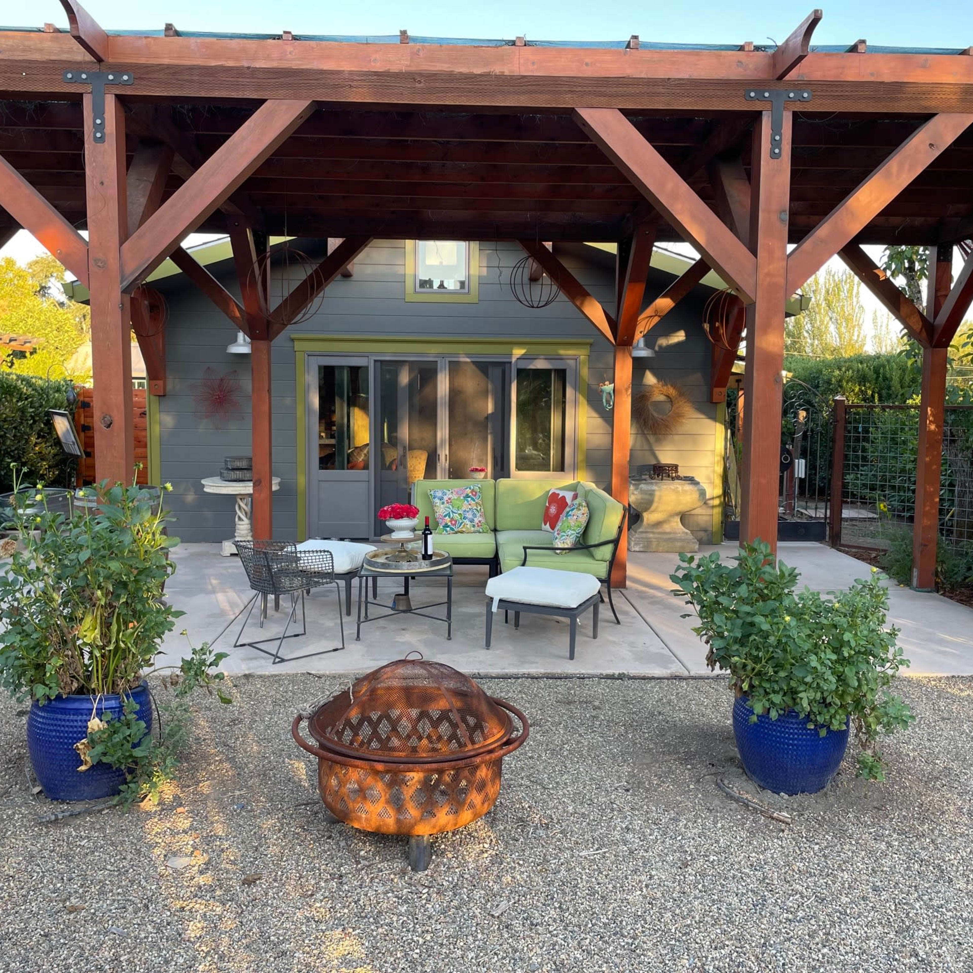 A patio area features a seating arrangement with green cushions, surrounded by potted plants and a fire pit, under a wooden pergola.