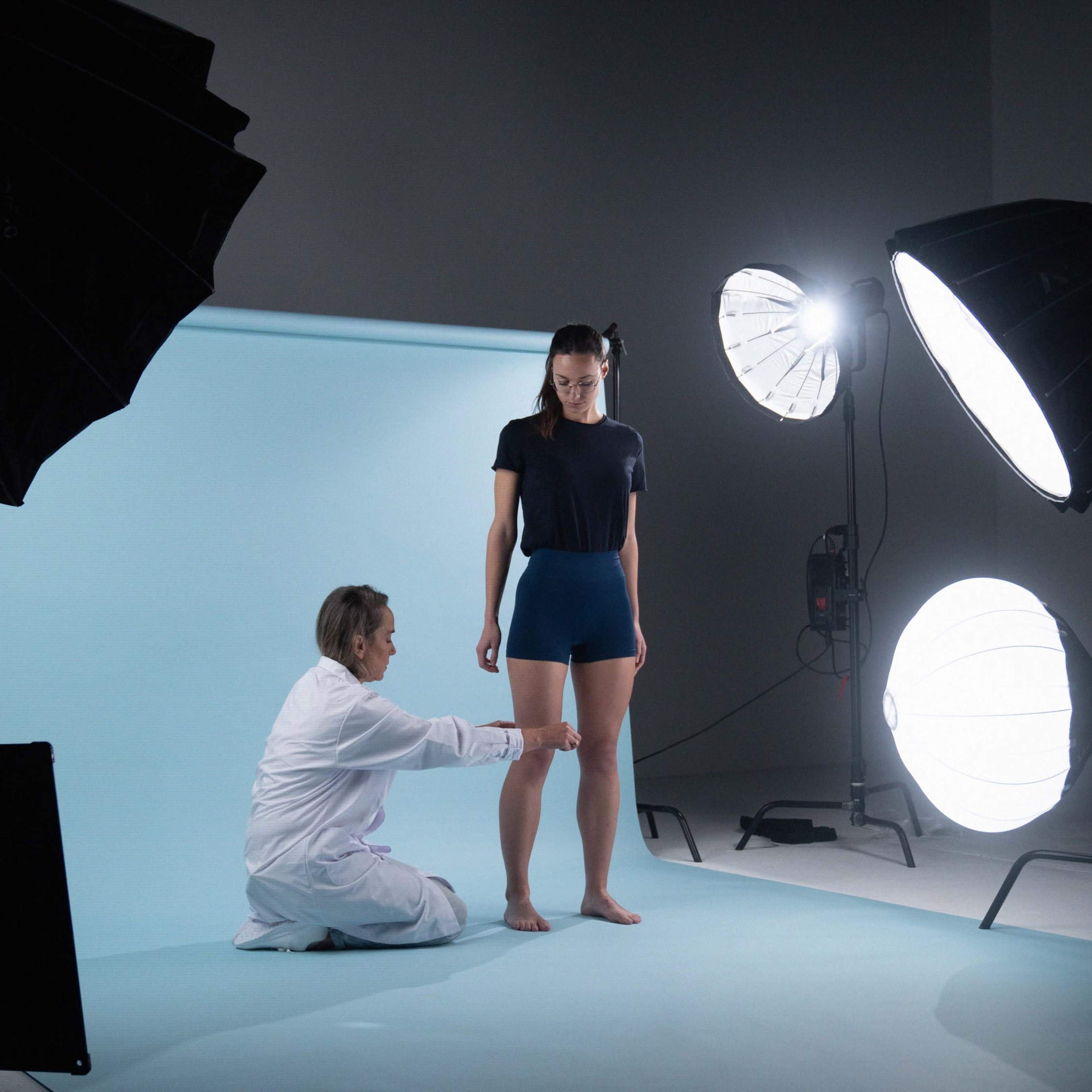 A photographer adjusts a model's pose in a studio setting with large softbox lights and a blue backdrop.