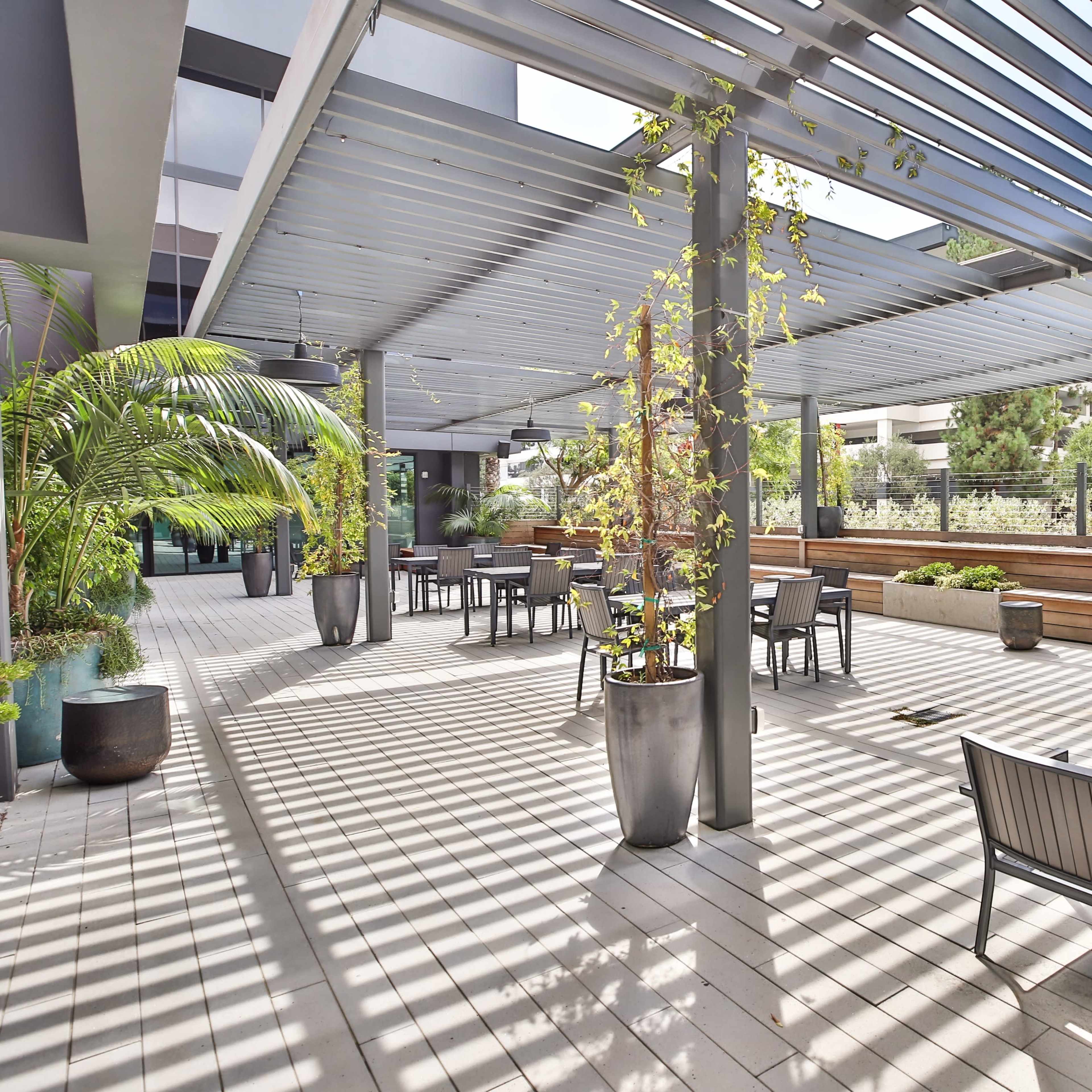 An outdoor patio area with shaded seating, planters, and wooden benches surrounded by greenery.