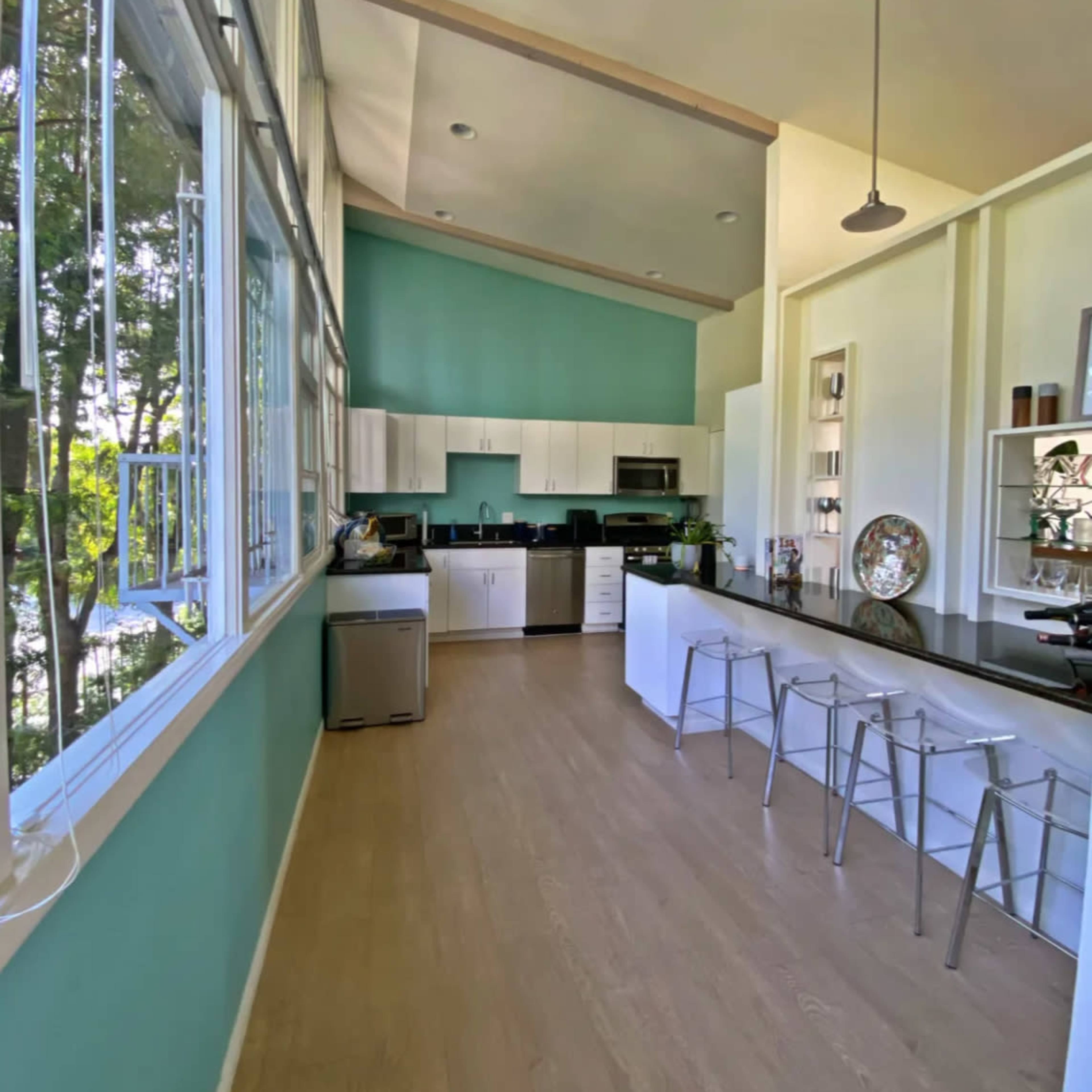 A modern kitchen features light-colored cabinets, a large central island with bar stools, and large windows that allow natural light to fill the space.