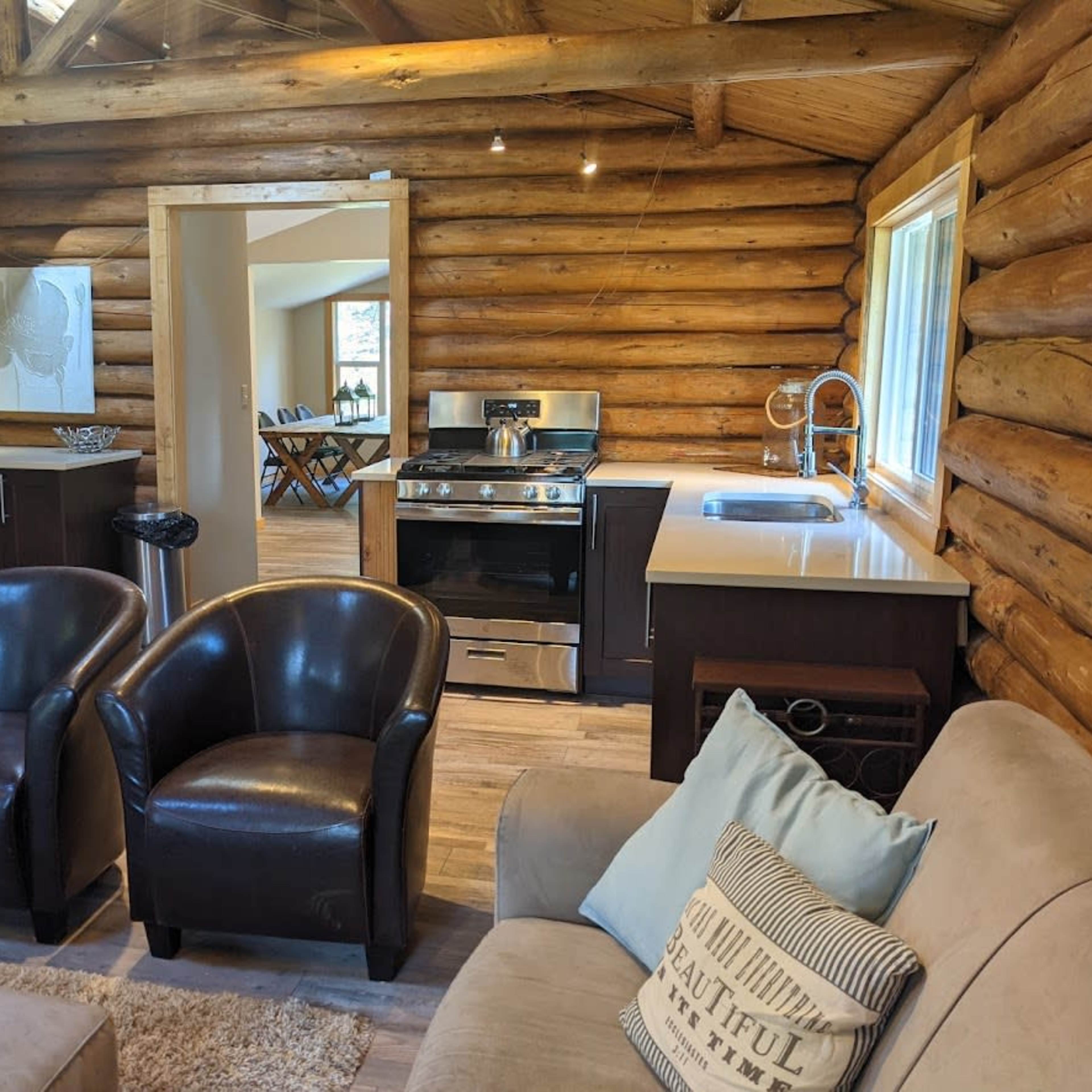 The interior of a log cabin features a kitchen with modern appliances, a dining area, and two leather chairs facing a sofa.