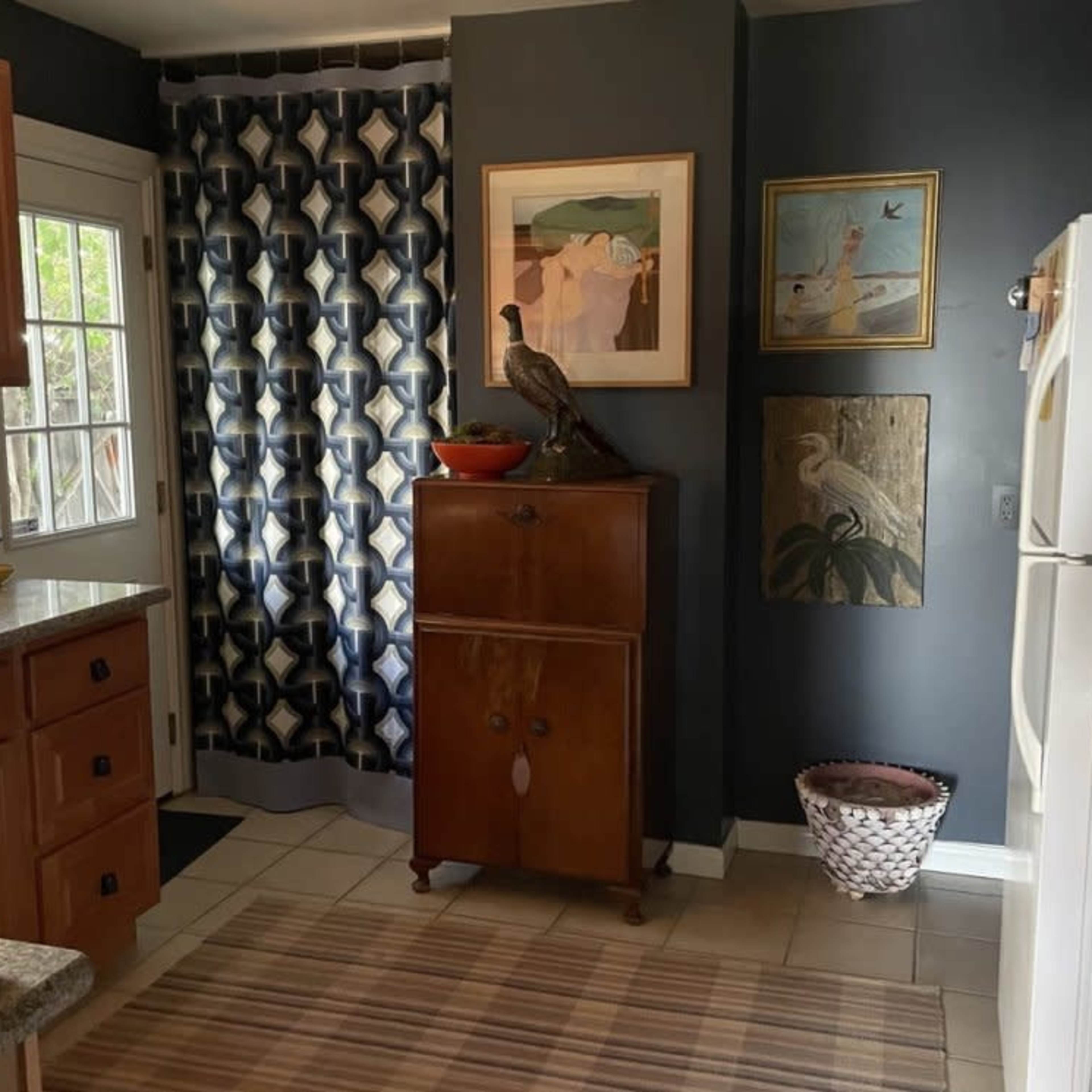 A kitchen with wooden cabinets, a decorative curtain, a vintage cabinet, and various wall art.