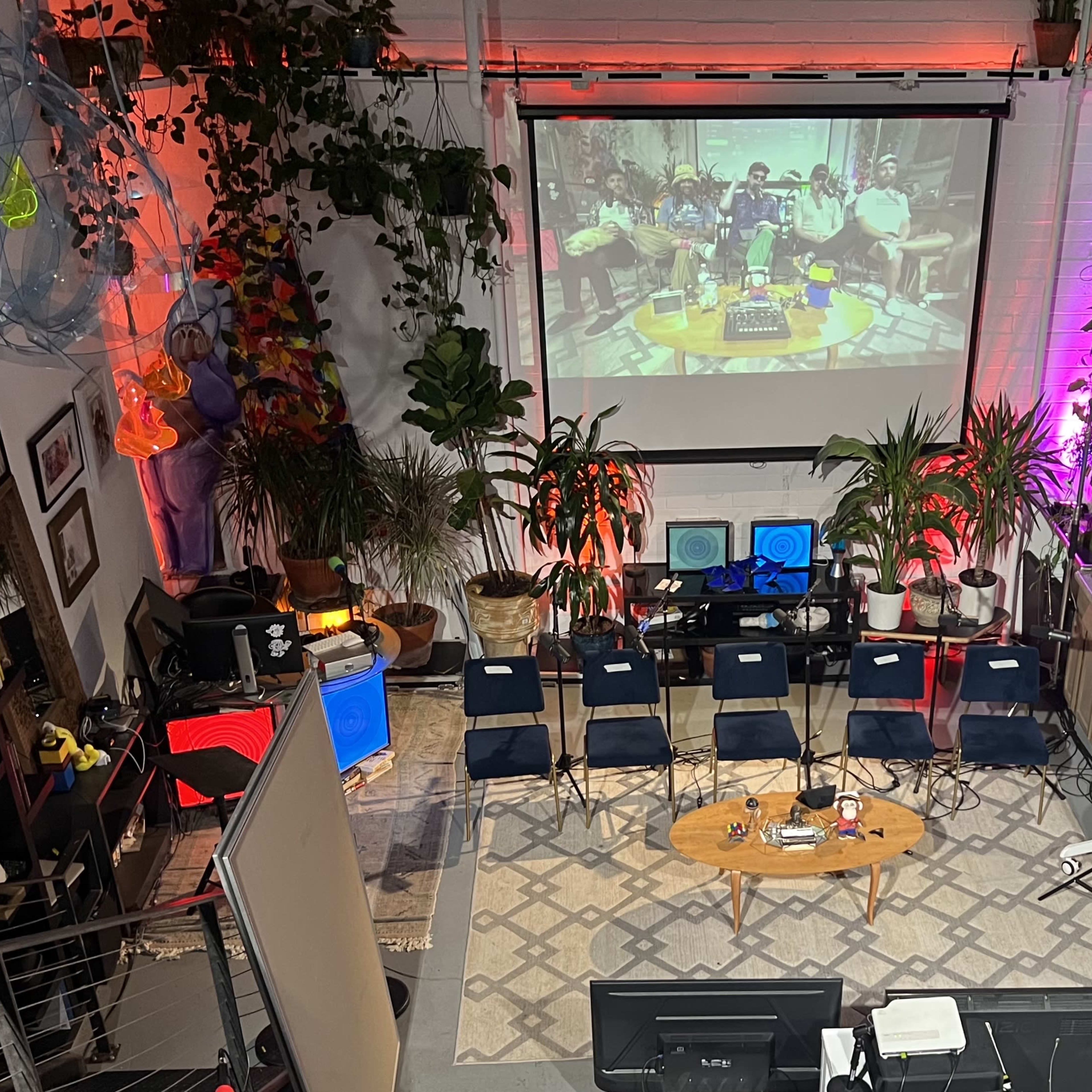 A vibrant indoor space set up with a large screen displaying a performance, surrounded by potted plants, chairs, and various audio equipment.