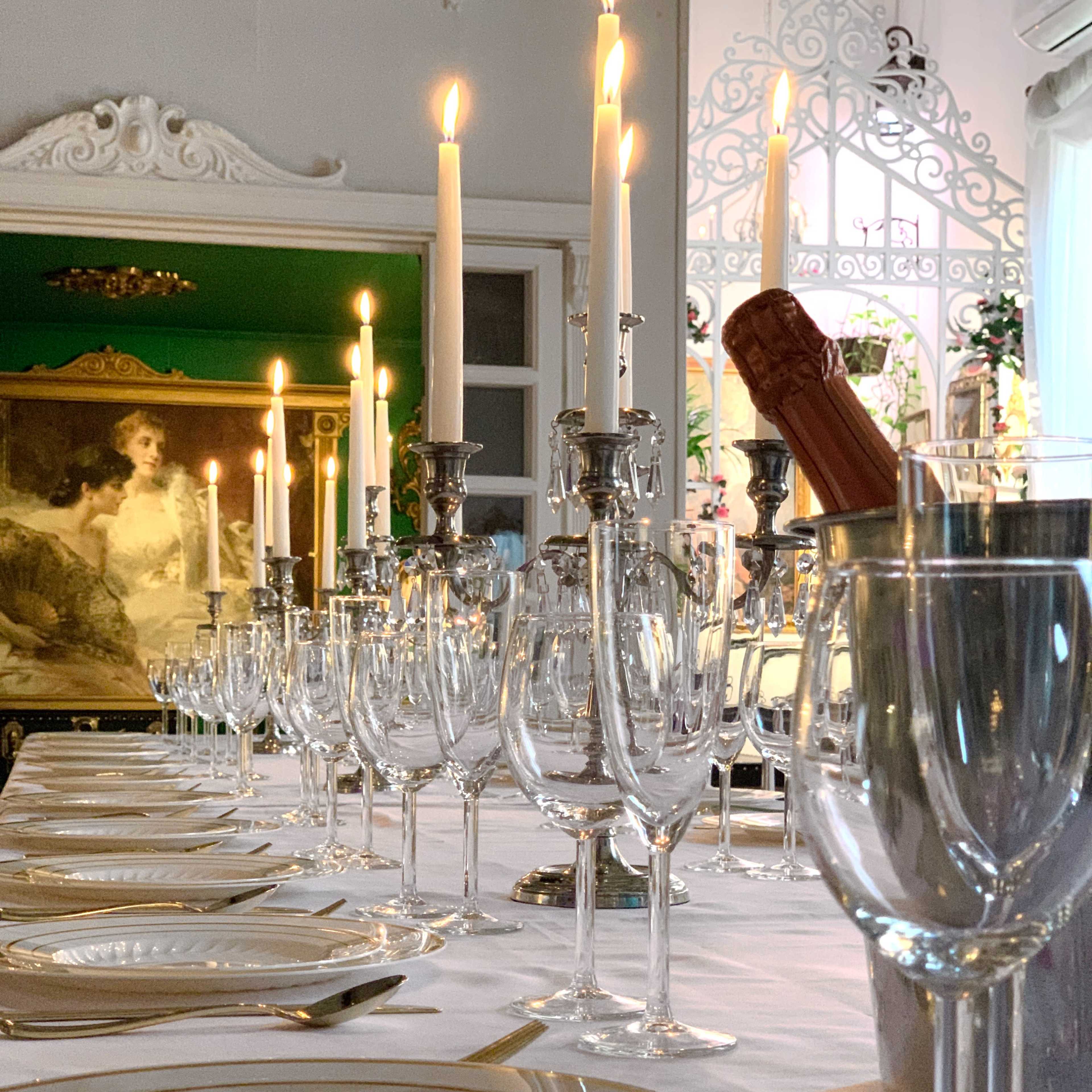 A long dining table is set with elegant glassware, polished silverware, and lit candles, with a painting visible in the background.