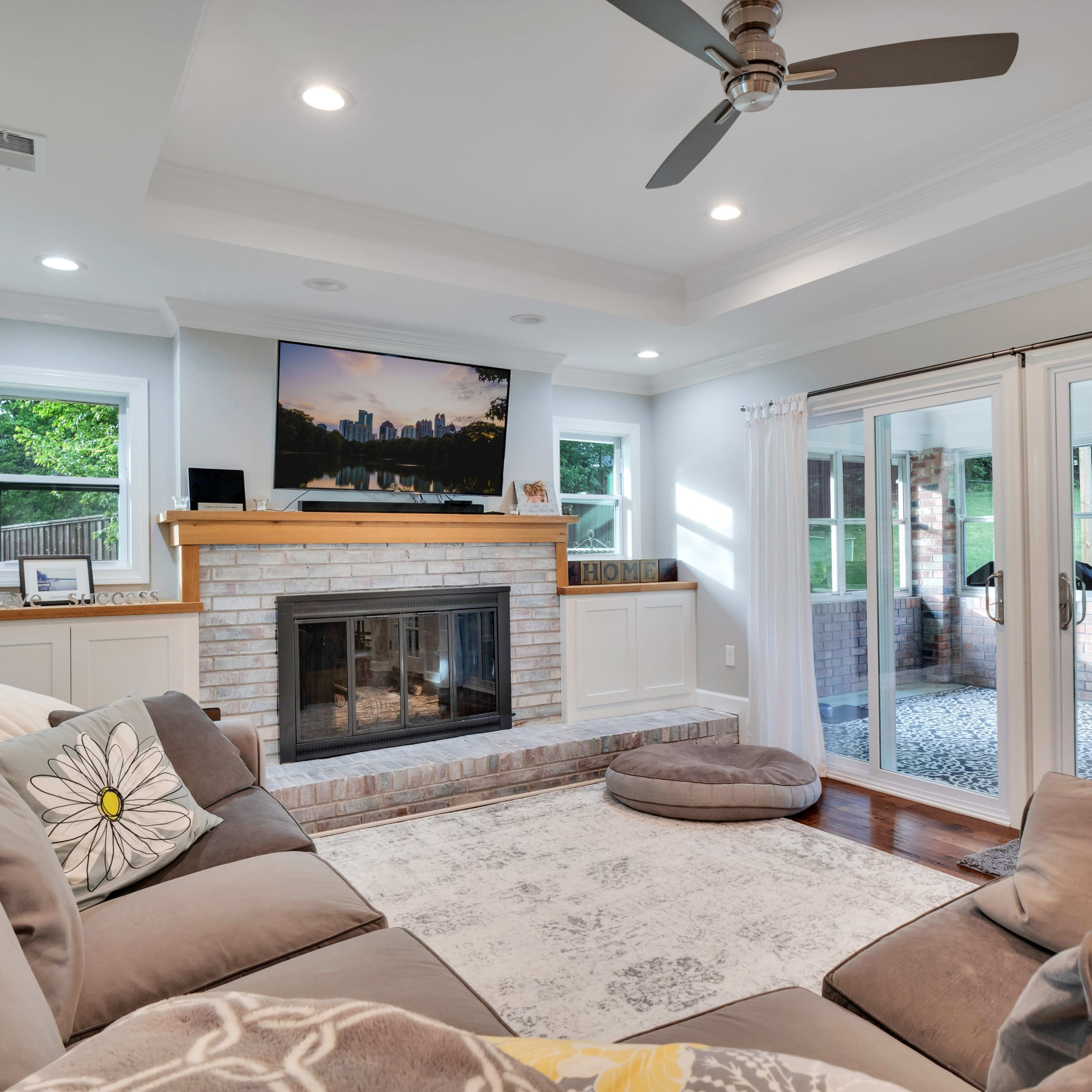 The living room features a cozy sectional sofa, a brick fireplace with a flat-screen TV above it, and large windows leading to an outdoor space.