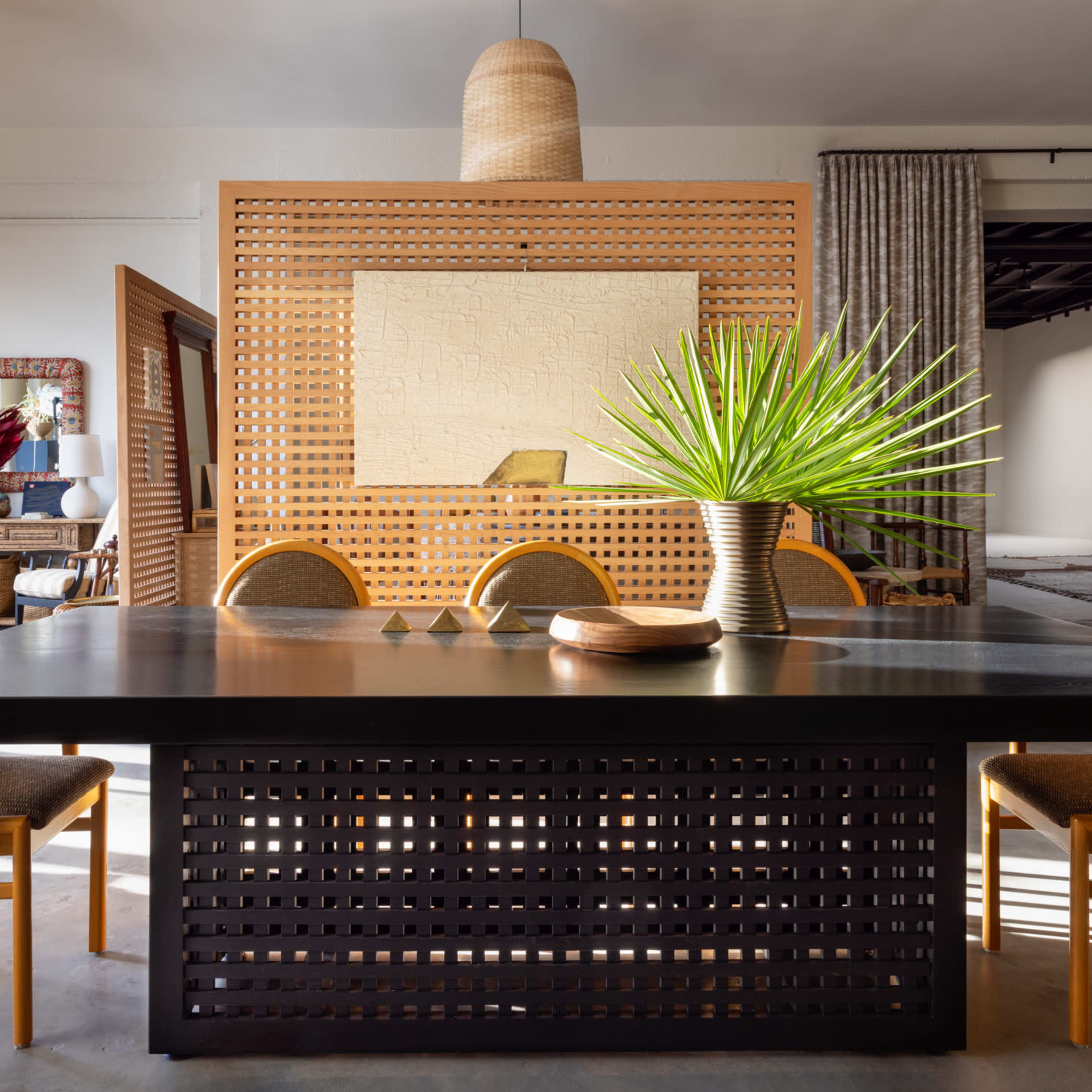 A large black table surrounded by six chairs is set in a well-lit dining area, featuring a decorative screen and a potted plant as the centerpiece.