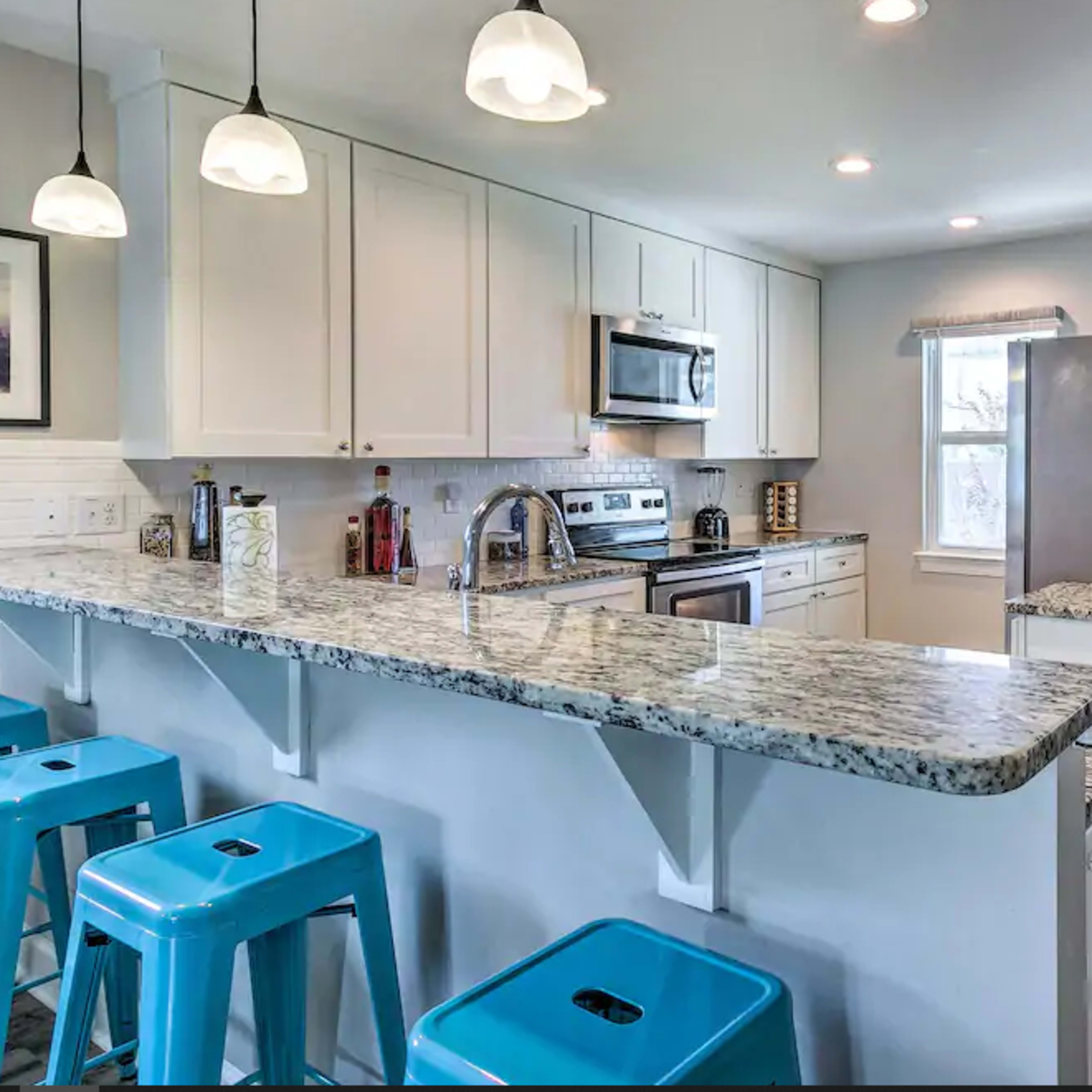 A modern kitchen with white cabinetry, a granite countertop, stainless steel appliances, and blue metal bar stools at a breakfast bar.