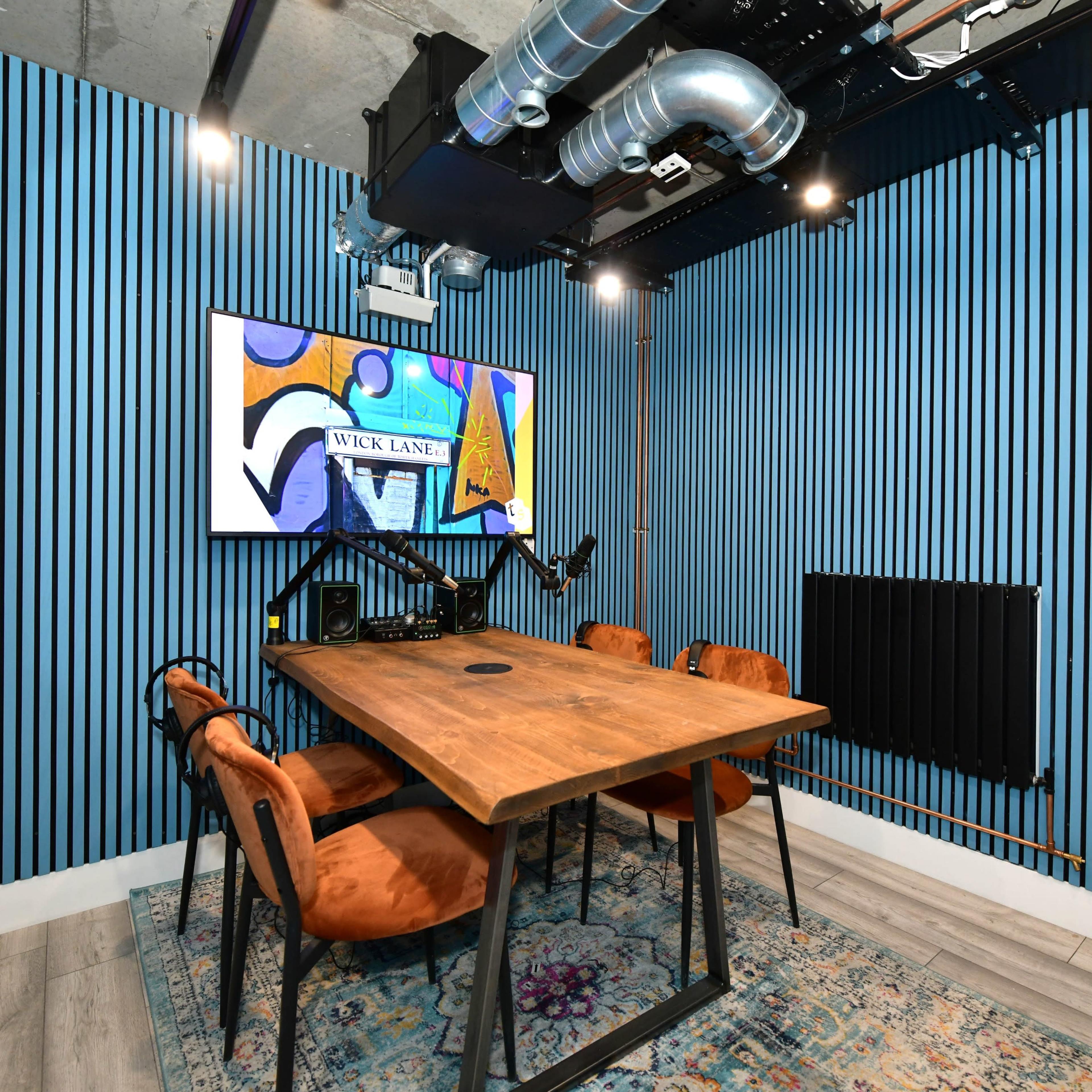 A small meeting room features a wooden table surrounded by four chairs, with a colorful artwork displayed on the wall and blue striped paneling.