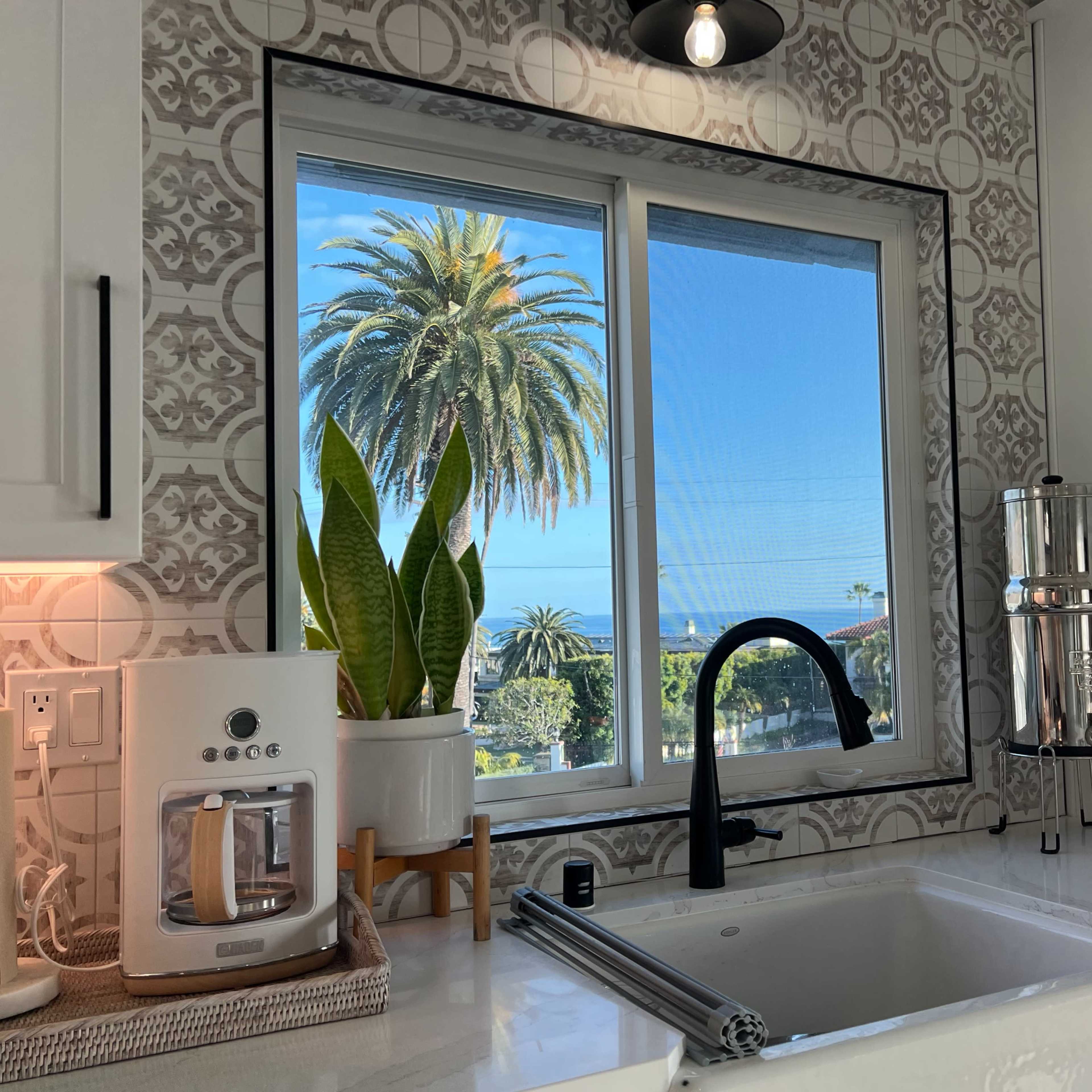 A kitchen with a modern sink, coffee maker, and a large window showcasing palm trees and a blue sky outside.