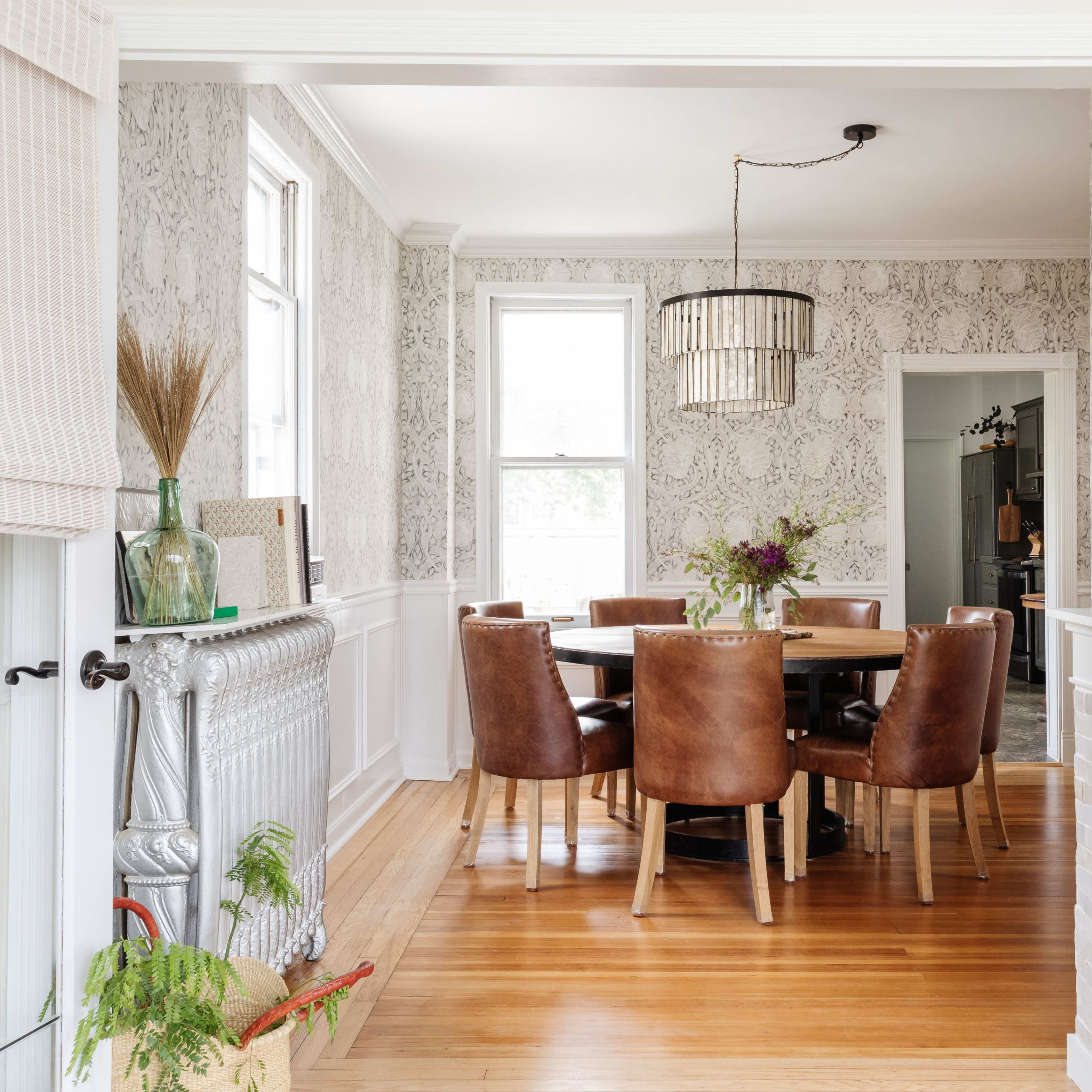 A dining room features a wooden table surrounded by brown leather chairs, with patterned wallpaper and a modern light fixture above.