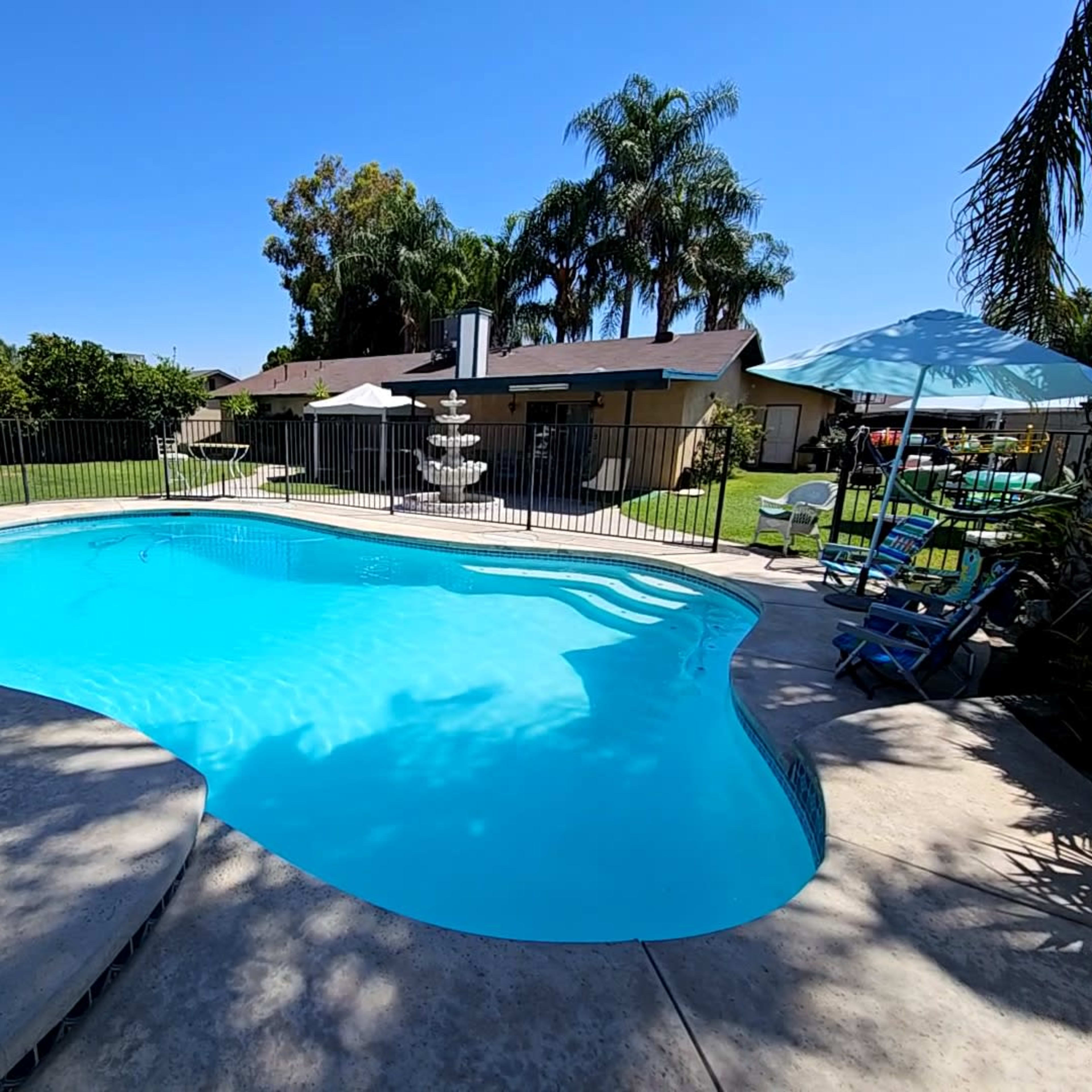 A clear swimming pool is surrounded by a concrete patio and a lush green backyard with palm trees and outdoor furniture.
