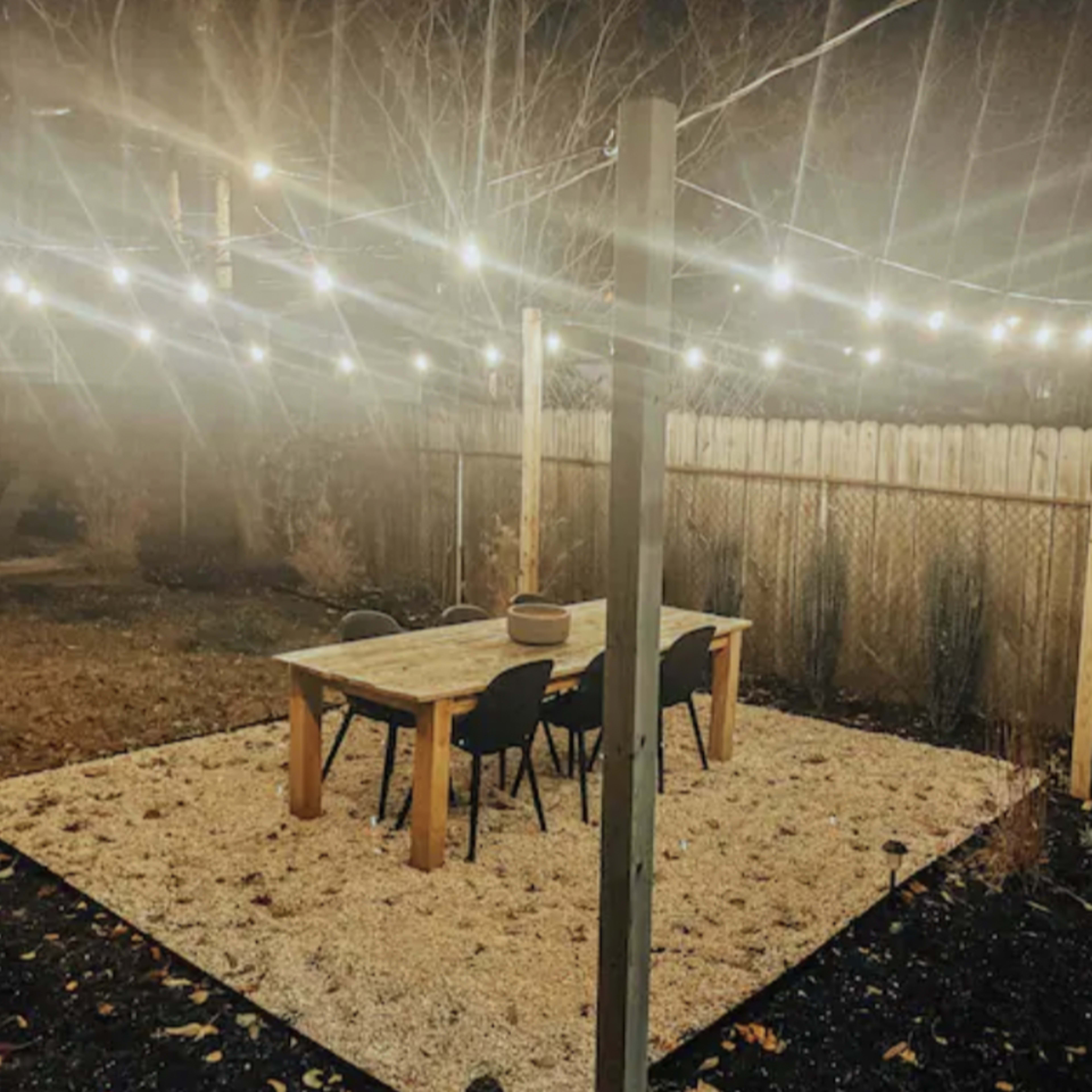 A patio area features a wooden table surrounded by chairs under string lights, with a gravel surface and a wooden fence in the background.