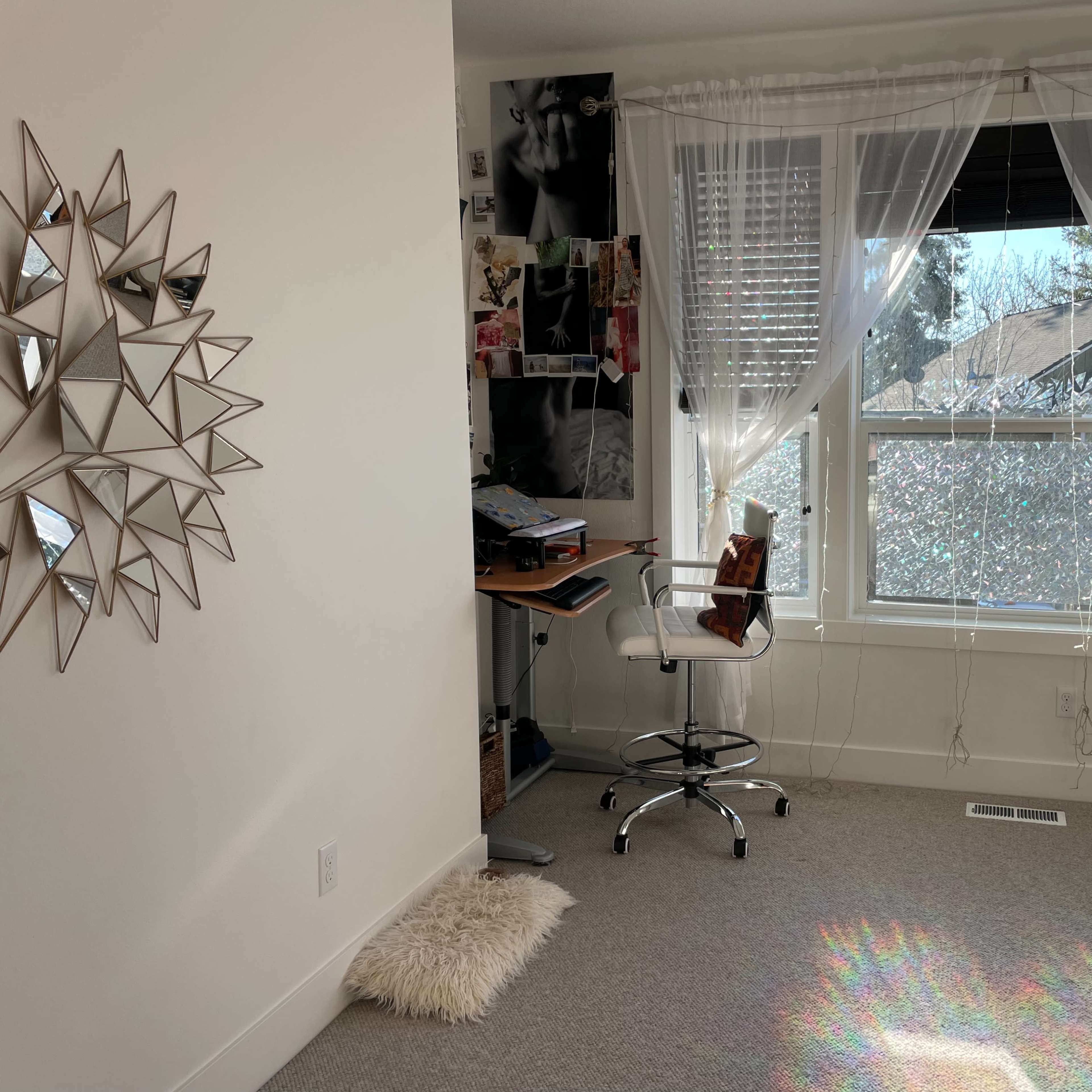 A bright room with a desk and chair positioned near a large window, adorned with light curtains, and featuring a decorative mirror on the wall.