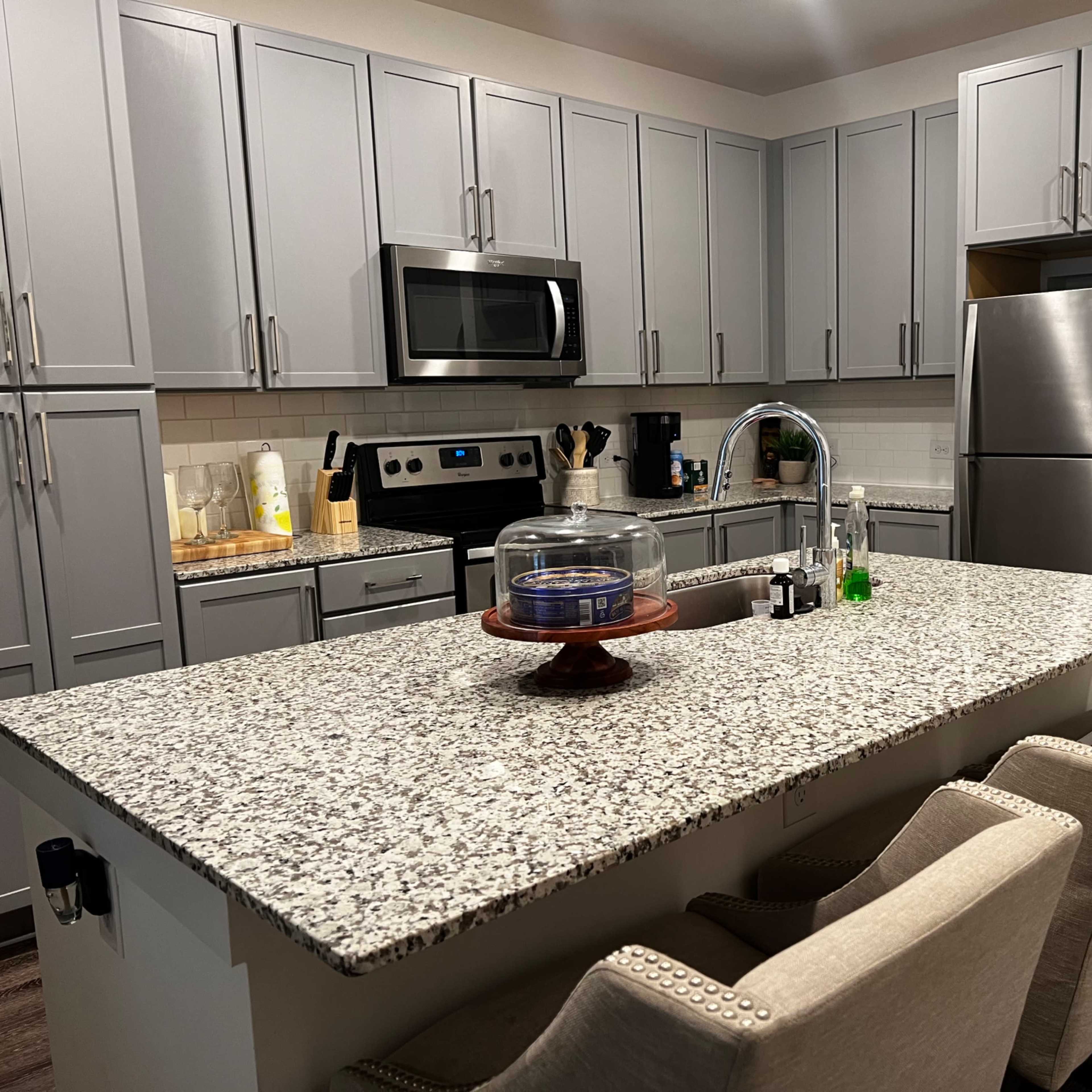 A modern kitchen with gray cabinets, a granite island, stainless steel appliances, and four upholstered bar stools.