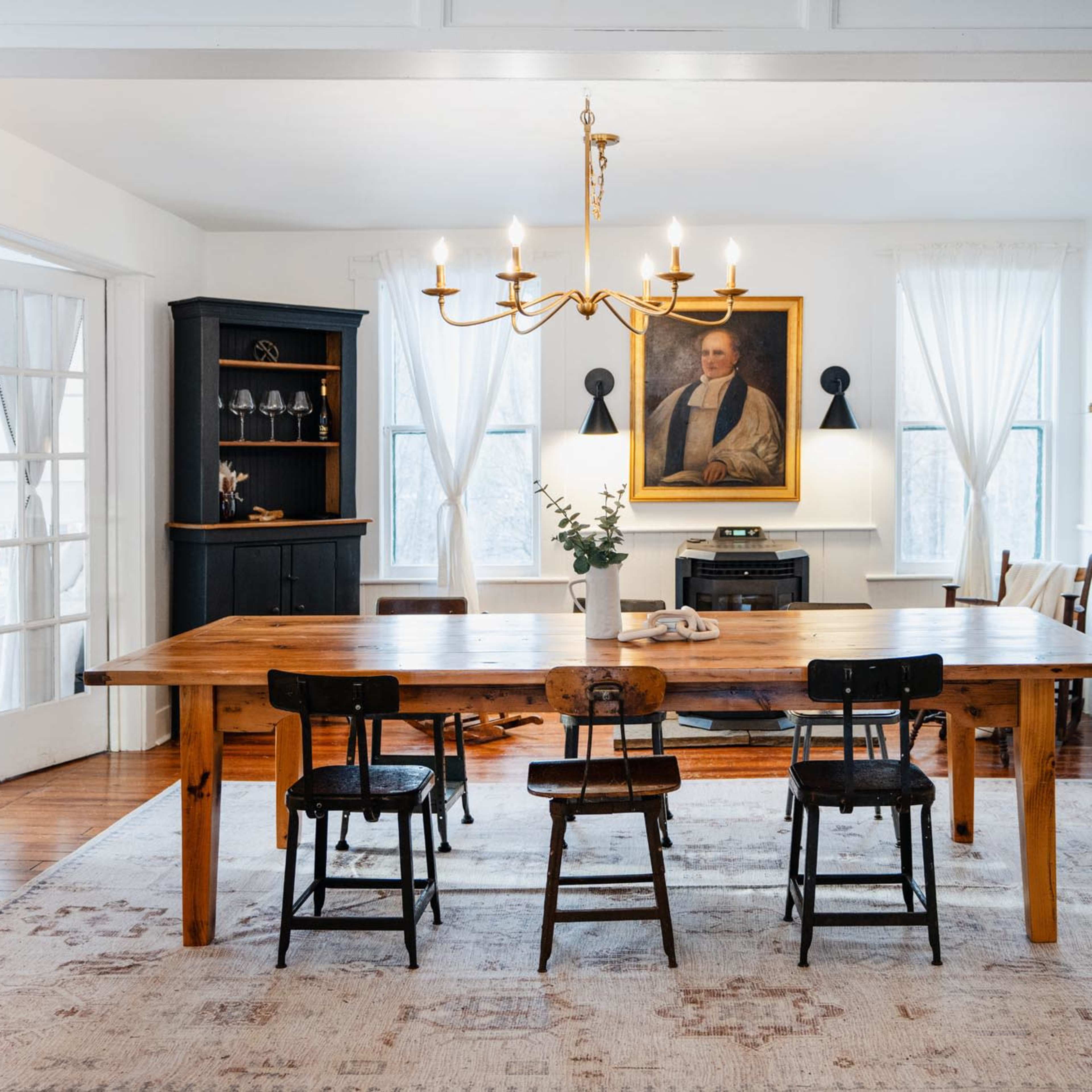 A dining room with a large wooden table surrounded by four chairs, framed by white walls and a portrait hanging above a black cabinet.